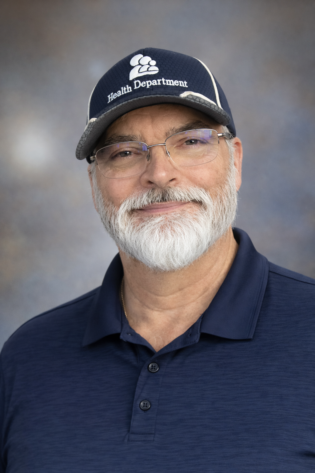 A smiling older man with a white beard and glasses, wearing a navy polo shirt and a navy cap with the words 'Health Department' and a logo on it, against a blurred gray backdrop.
