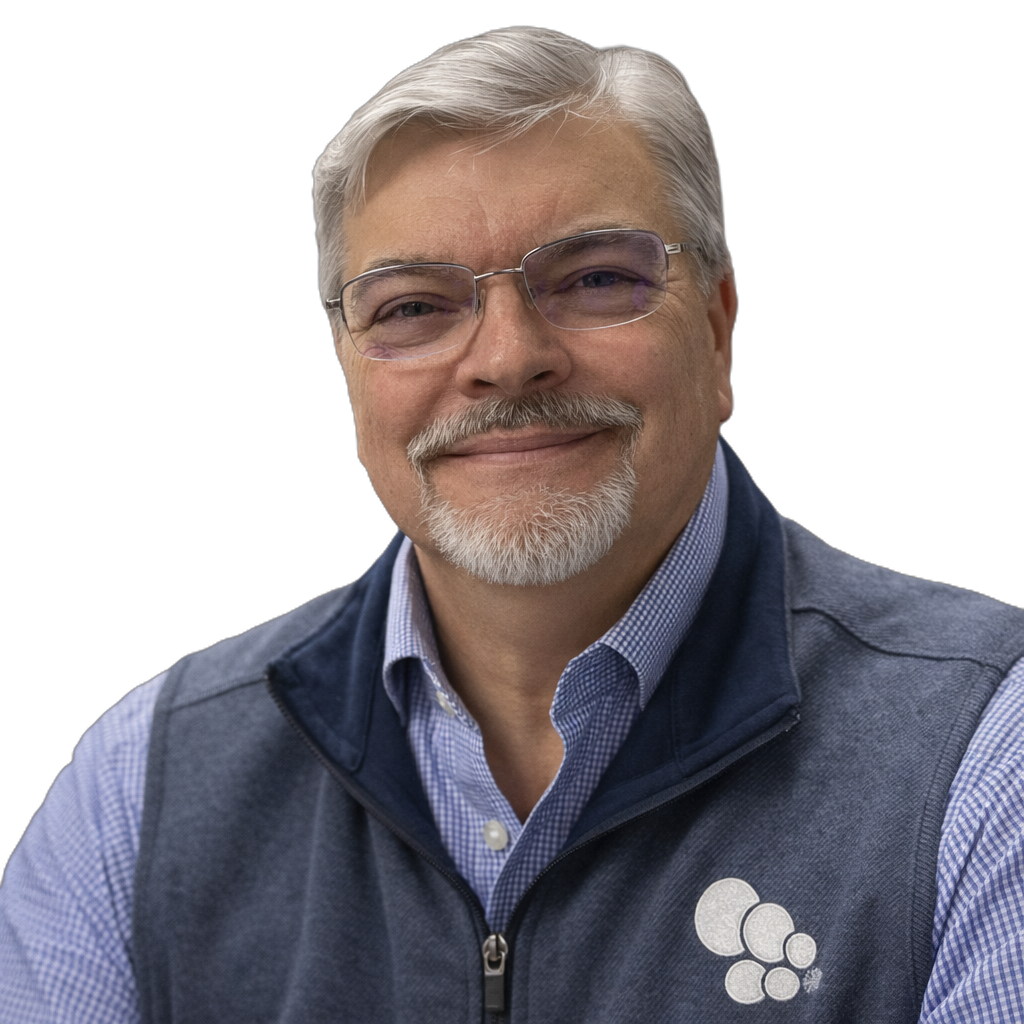 A middle-aged man with gray hair, a mustache, and glasses, smiling, wearing a blue checkered shirt and a dark blue vest with a circular logo on it, against a black background.