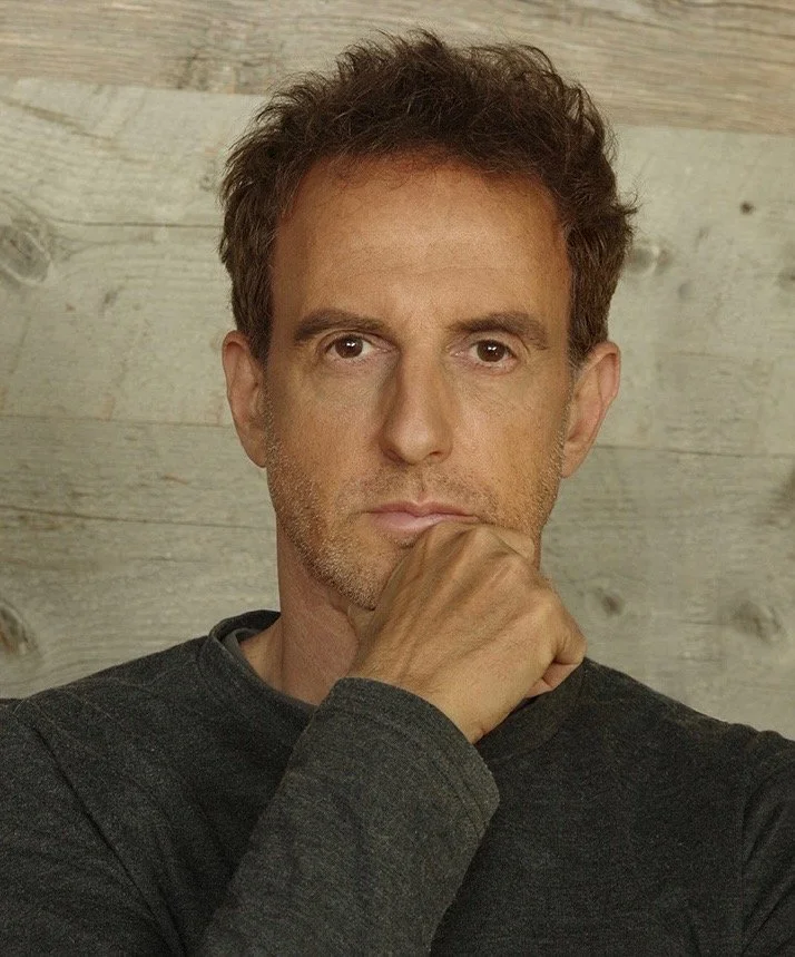 A middle-aged man with short brown hair and a serious expression, resting his chin on his hand, wearing a dark long-sleeve shirt, against a light wooden background.
