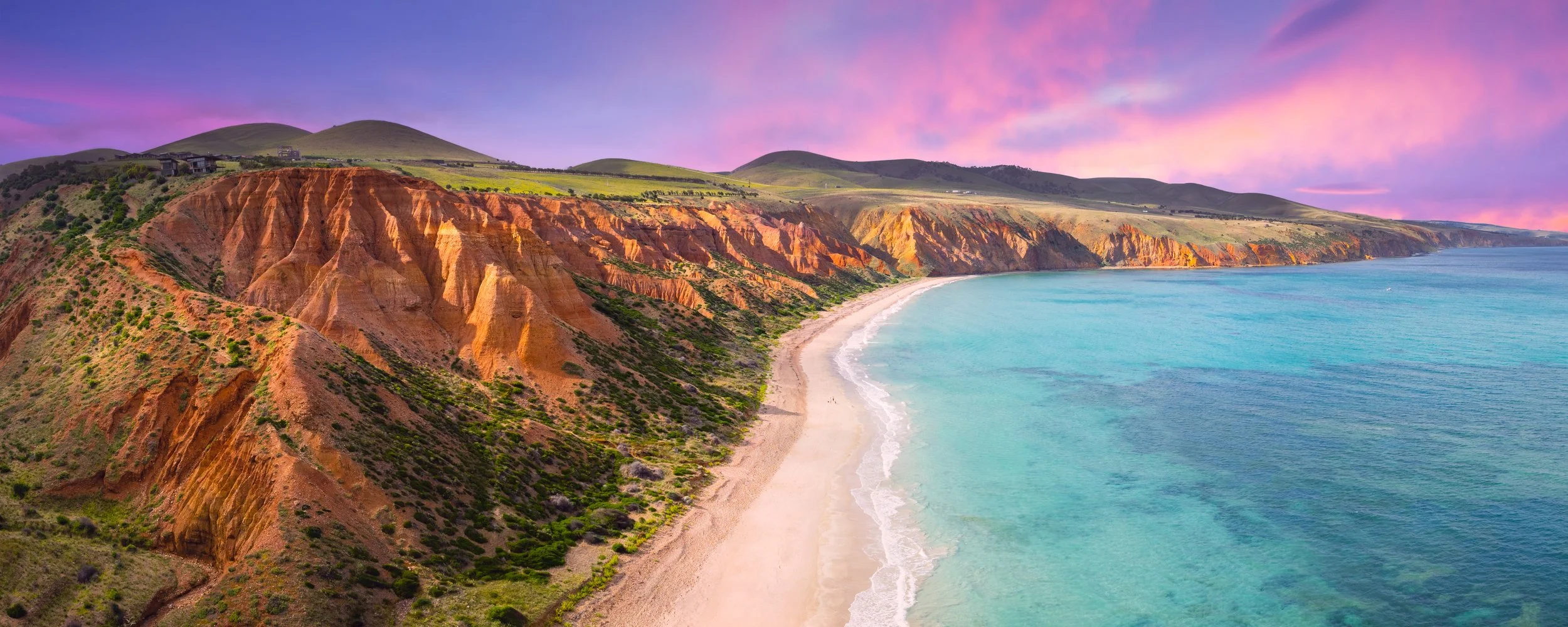 Colorful coastal landscape with red and orange cliffs, sandy beach, and turquoise ocean under a pink and purple sky.