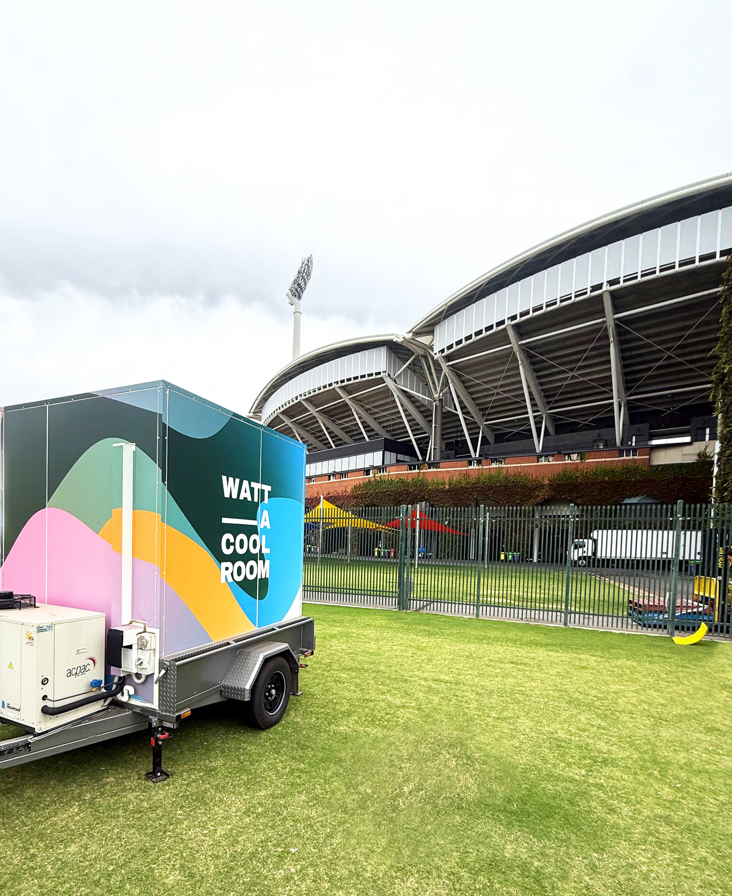 A man-made structure resembling a stadium with large curved roof panels, viewed behind a fenced grassy area. In the foreground, there is a colorful mobile unit with the words 'WATT A COOL ROOM' on it and some outdoor equipment, set against an overcast sky.