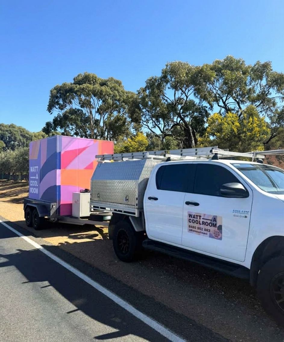 A white utility truck with a ladder on top and business signage on the side, parked next to a colorful mobile cooling or refrigeration unit trailer, on a roadside with trees and blue sky in the background.