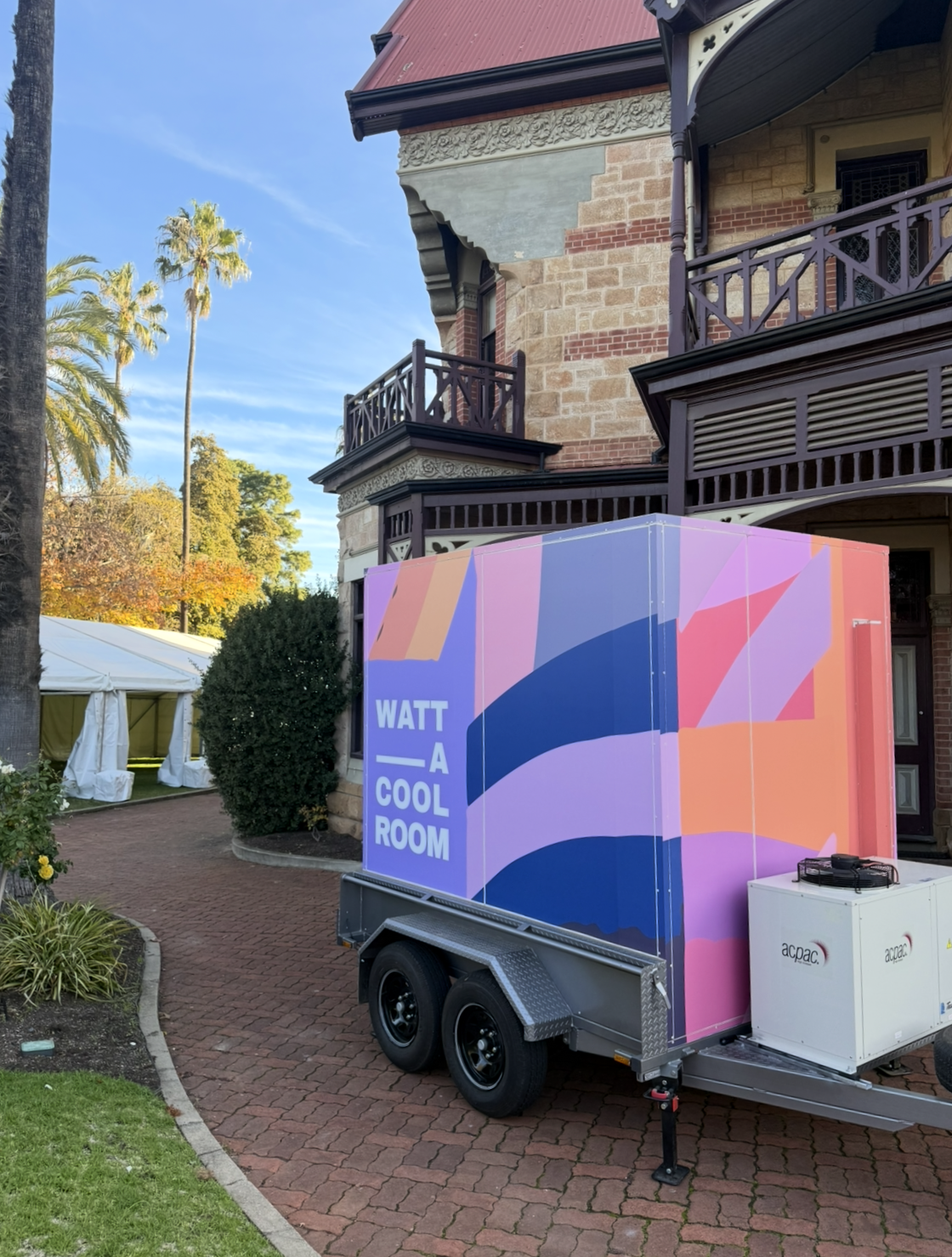 A colourful mobile cool room with the text 'WATT A COOL ROOM' displayed on the side in front of a historic building with ornate architectural details, a red-tiled roof, and a surrounding garden with trees and a tent.