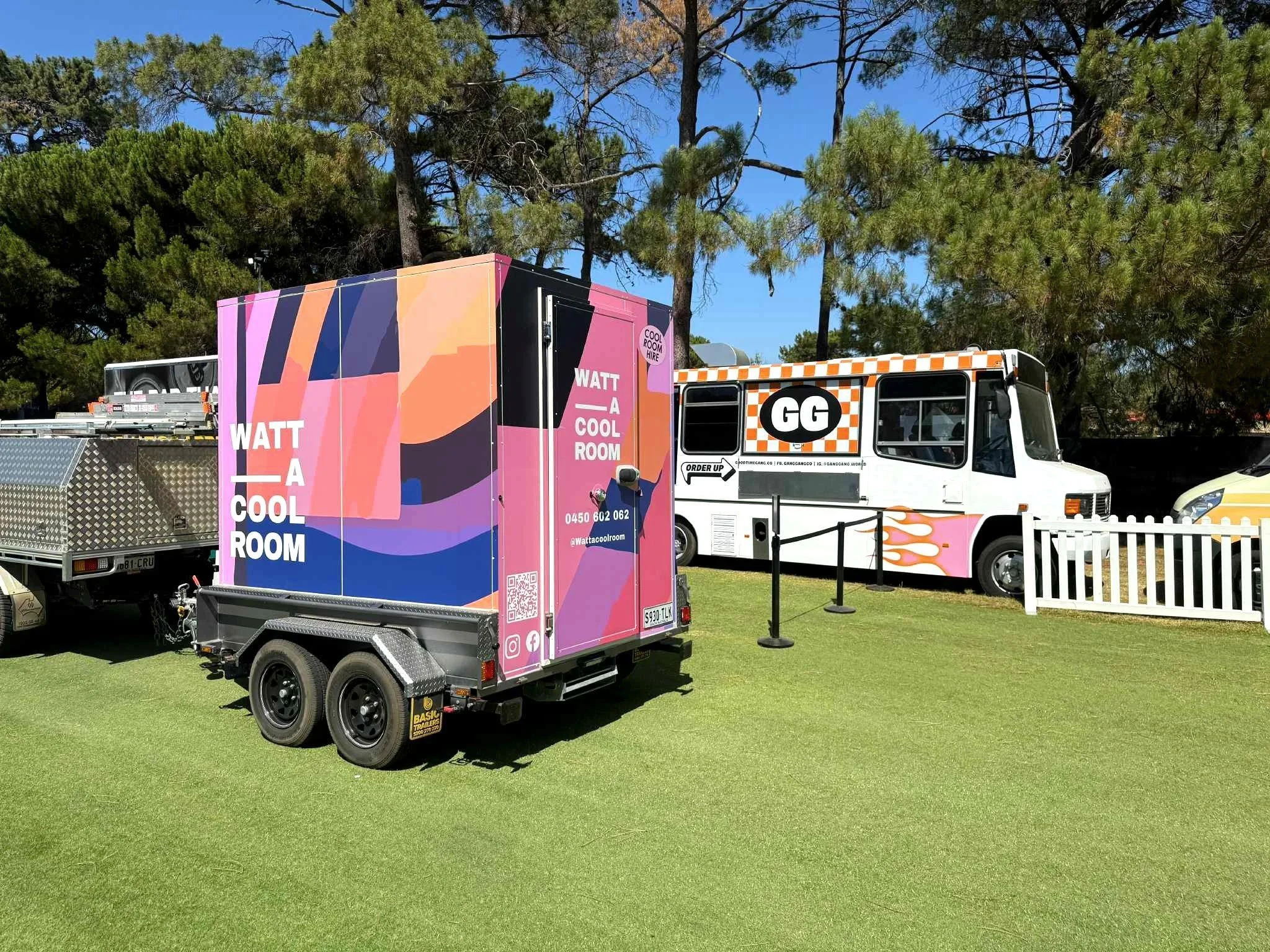 A mobile cool room with colourful artwork sitting on a green grass field under tall trees with a clear blue sky.