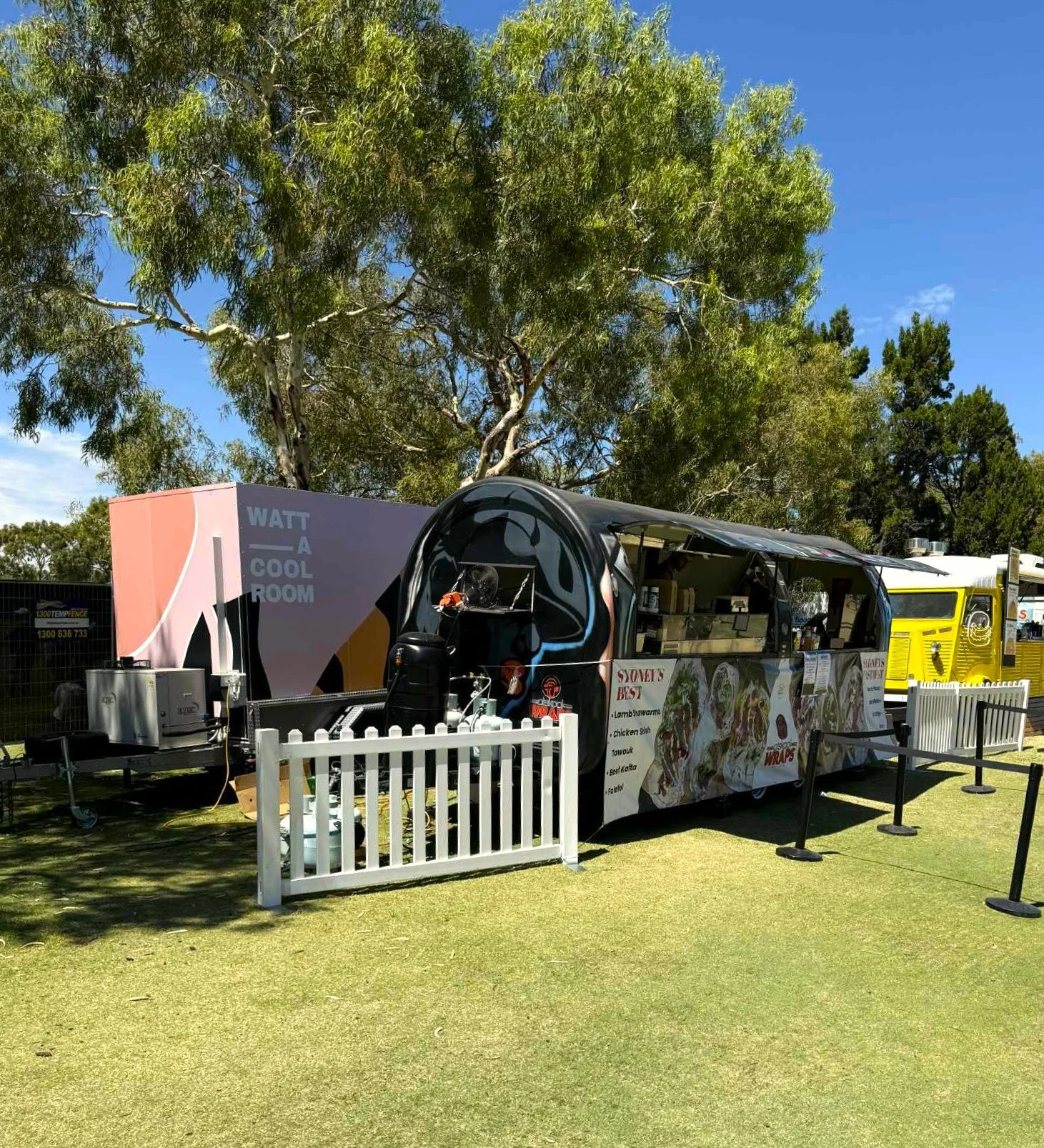 A mobile cool room and food truck parked near green grass with a large tree in the background. The truck has menu signs and a white picket fence in front.