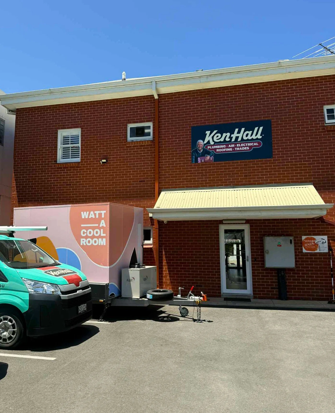 Brick building with sign promoting Ken Hall plumbing, air, electrical, roofing, trades services, and a delivery truck in the parking lot.