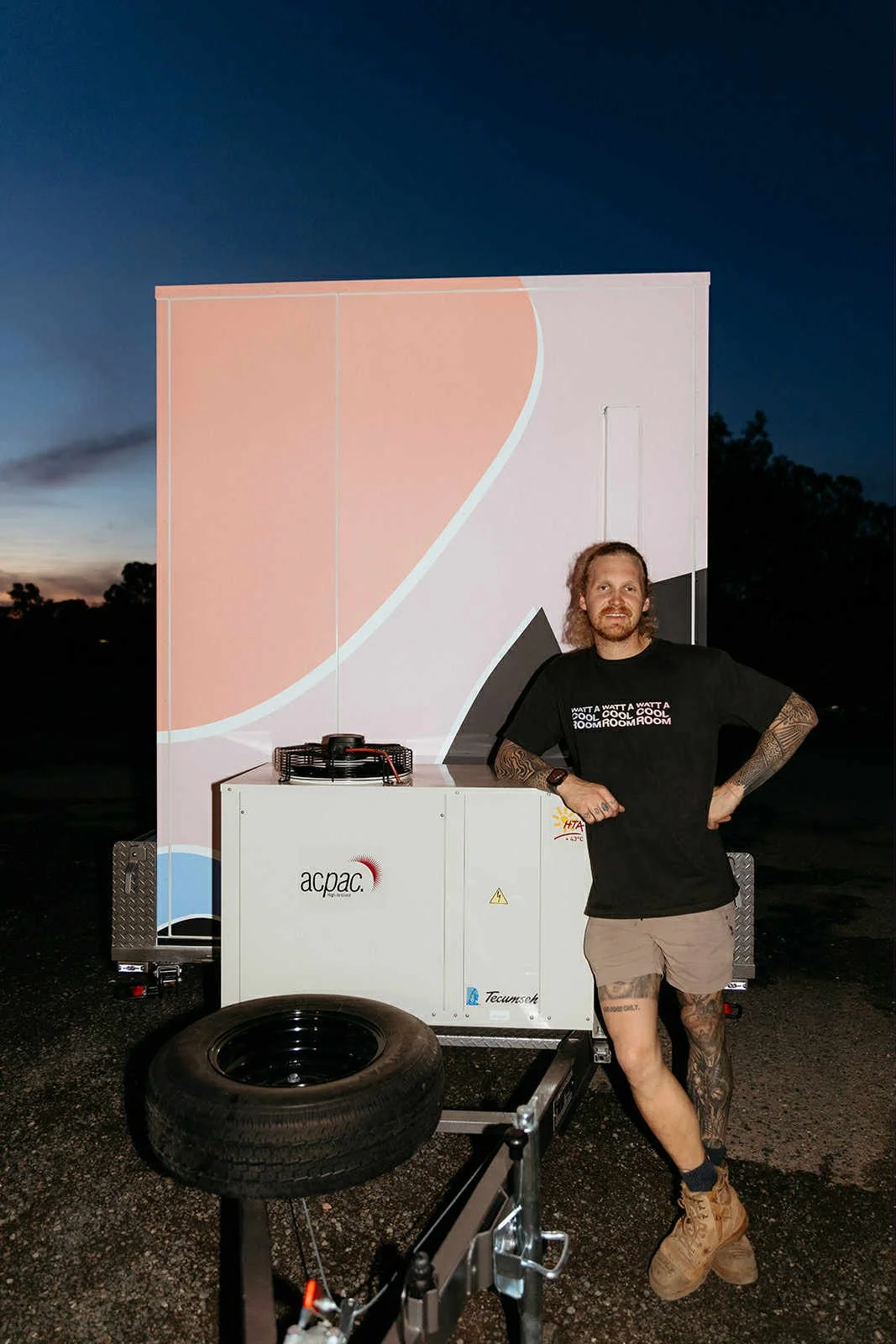 A man with long hair, tattoos, and wearing a black t-shirt and beige shorts, standing next to a mobile cool room outdoors at dusk.