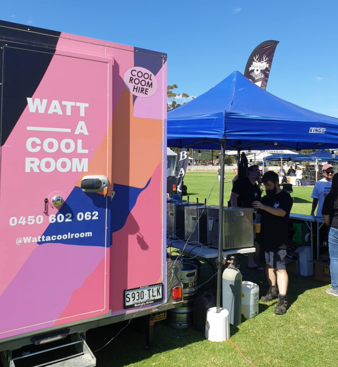 A colourful mobile cool room with the words "WATT A COOL ROOM" and contact information on its side, adjacent to a blue canopy tent where people are gathered at an outdoor event on a sunny day.