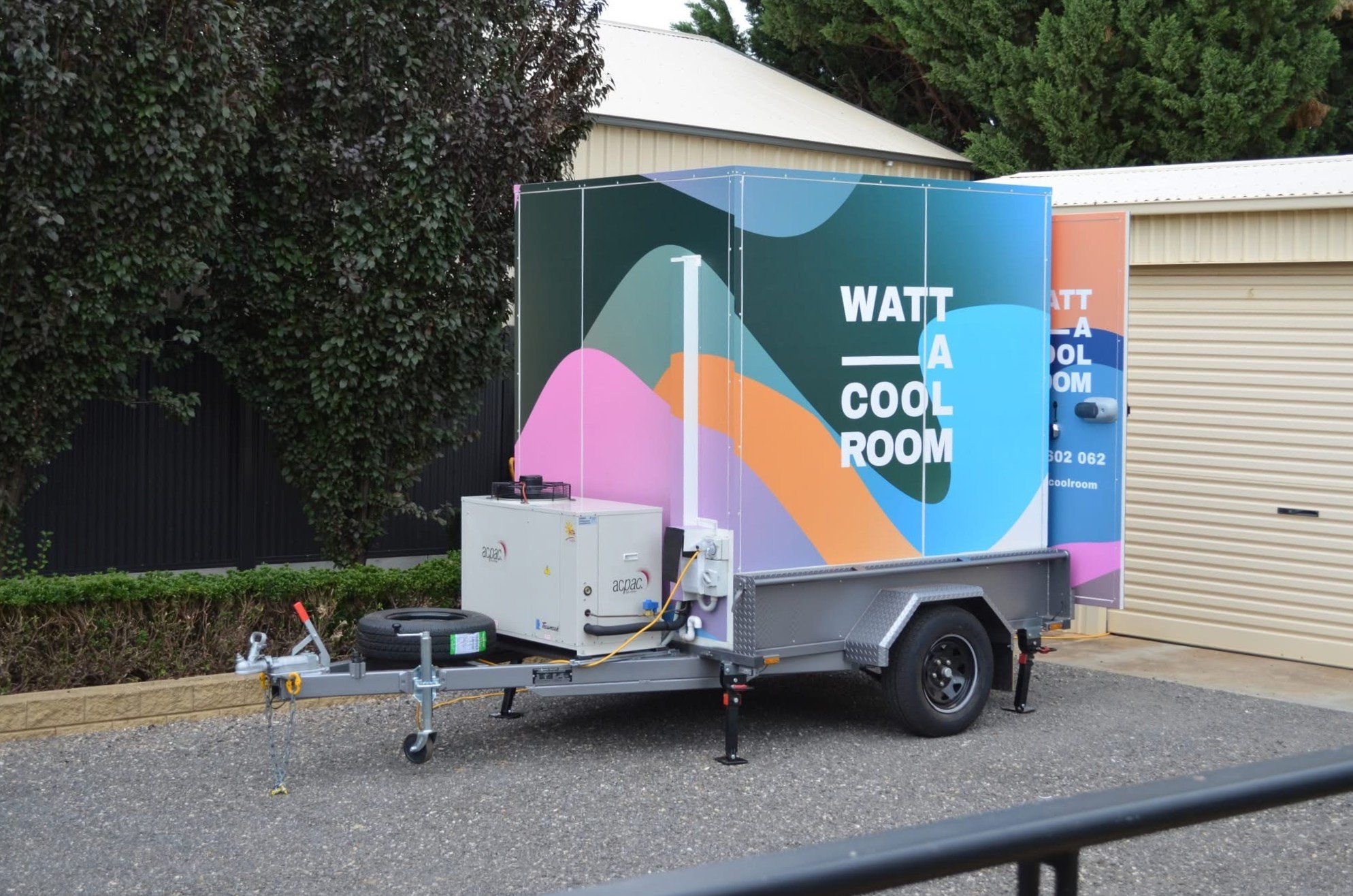 Mobile cooling unit with colorful branding promoting a cool room product, parked on a gravel surface next to a building, with greenery and trees in the background.