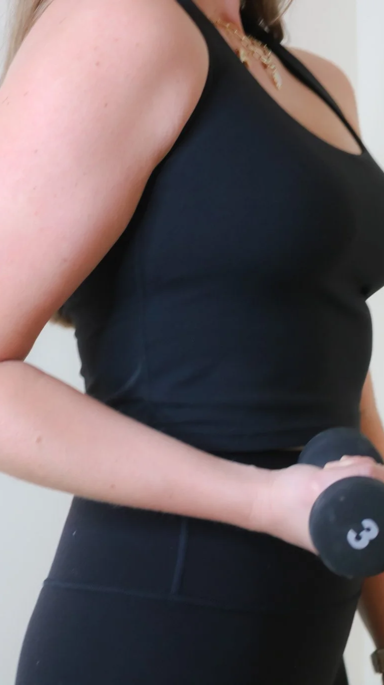 A woman holding a small dumbbell while wearing a black workout top and black leggings.