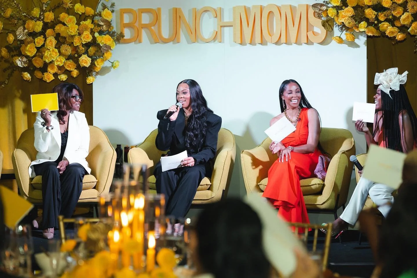 Four women seated on yellow chairs on a stage at the 'Brunch Moms' event, smiling and engaging in conversation. The backdrop features large yellow flowers and a sign that reads 'BRUNCH MOMS'. One woman is holding a microphone, and others are holding 