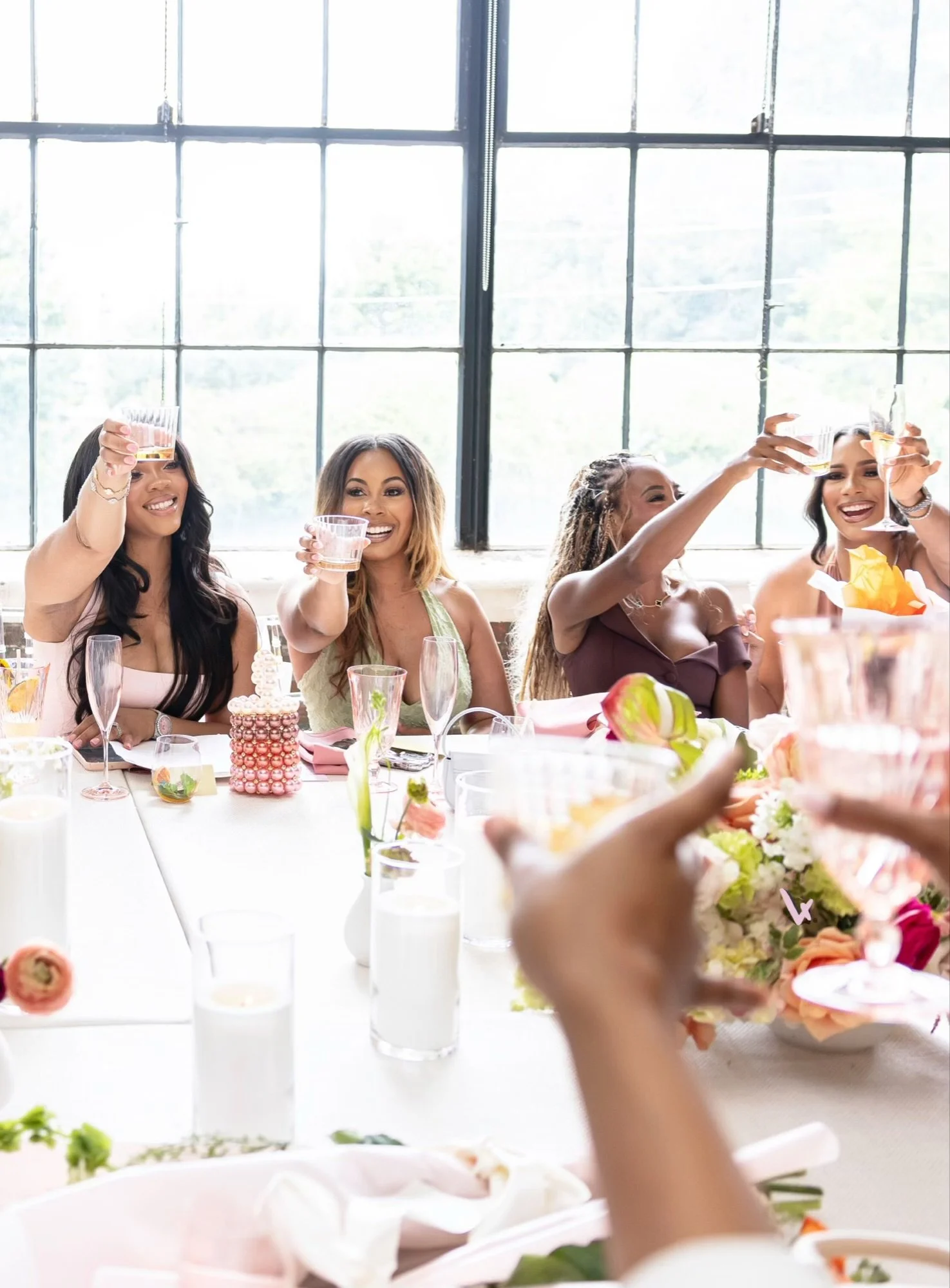 A group of women celebrating at a brunch or wedding event, raising glasses in a toast near a large window with a bright background.