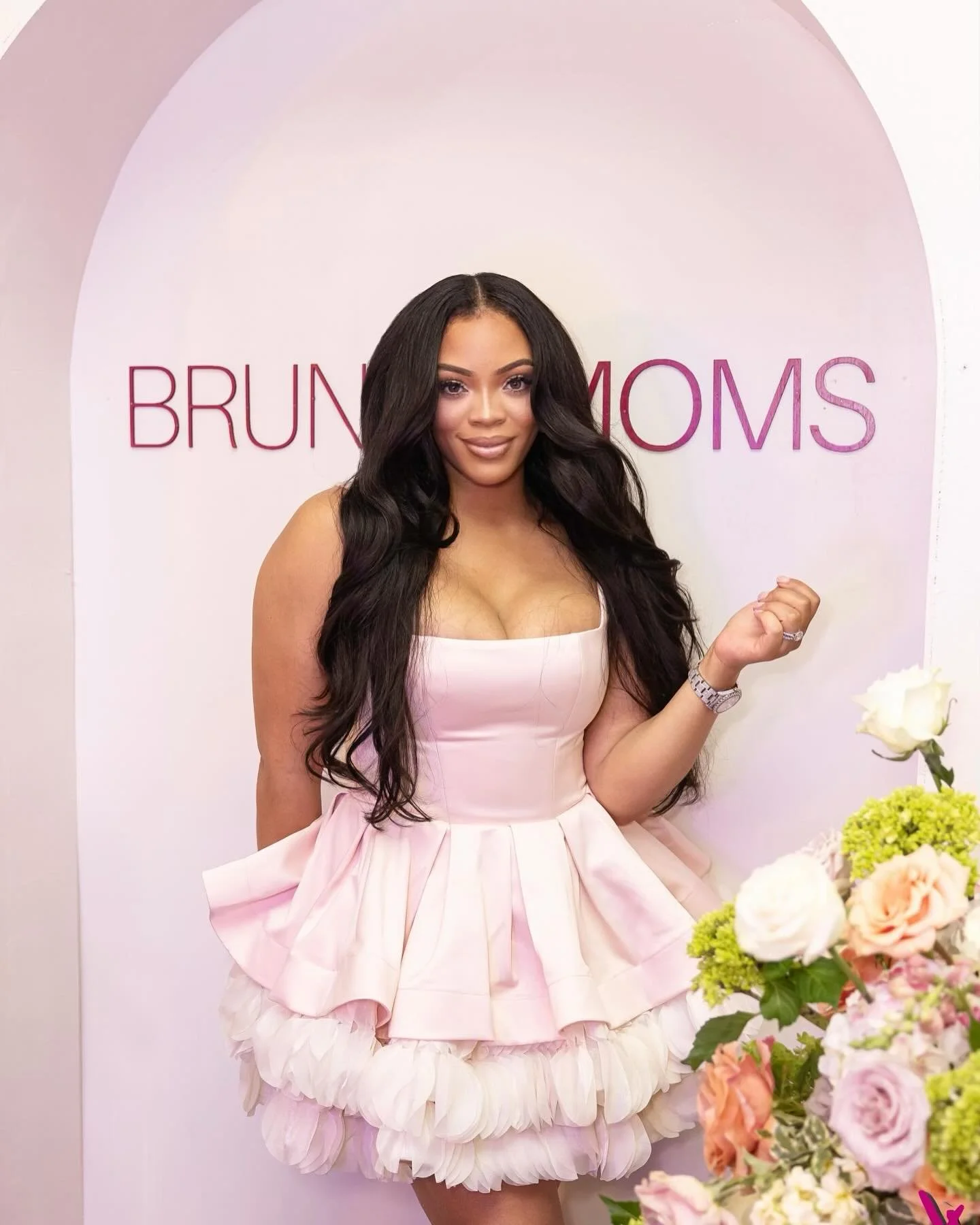 A woman in a pink dress standing in front of a sign that reads 'Brunch MOMS' with flowers nearby.