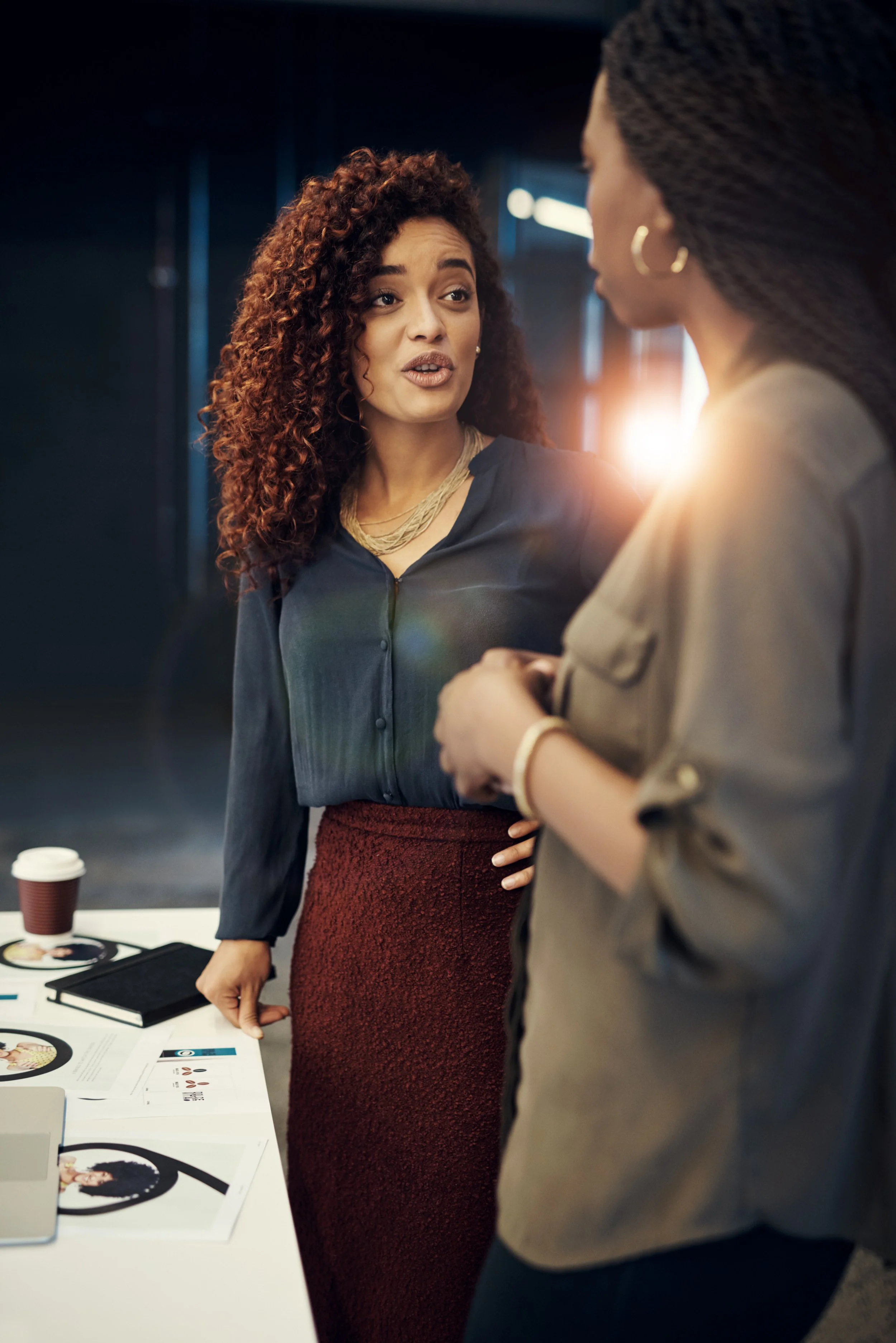 Two women having a conversation at a business meeting or conference, with documents and a coffee cup on the table.