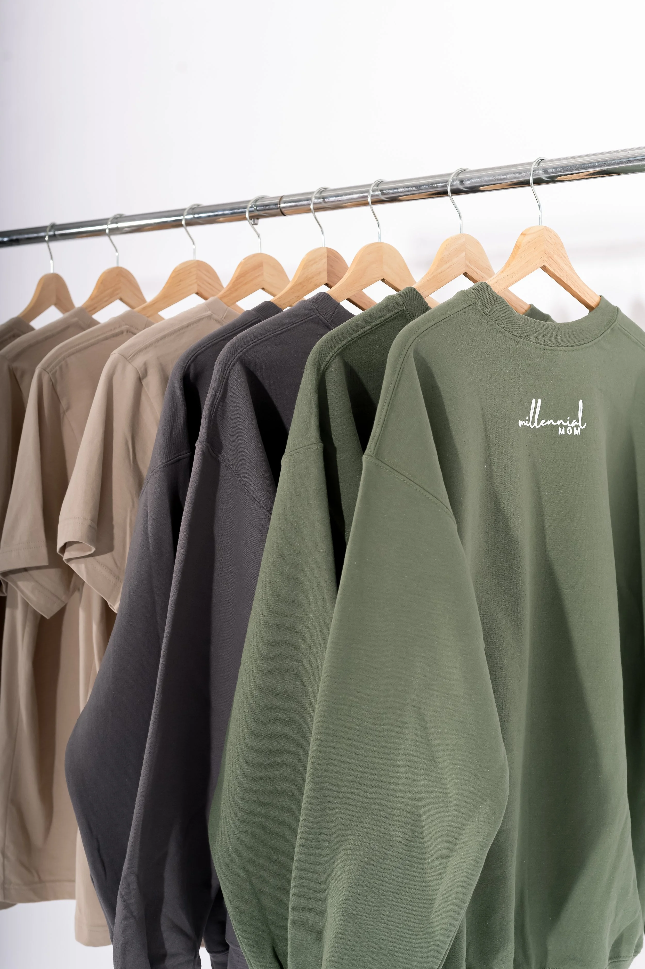 A row of hanging t-shirts in beige, black, and green colors, with the green shirt displaying the text 'millennial MOM' on the front.