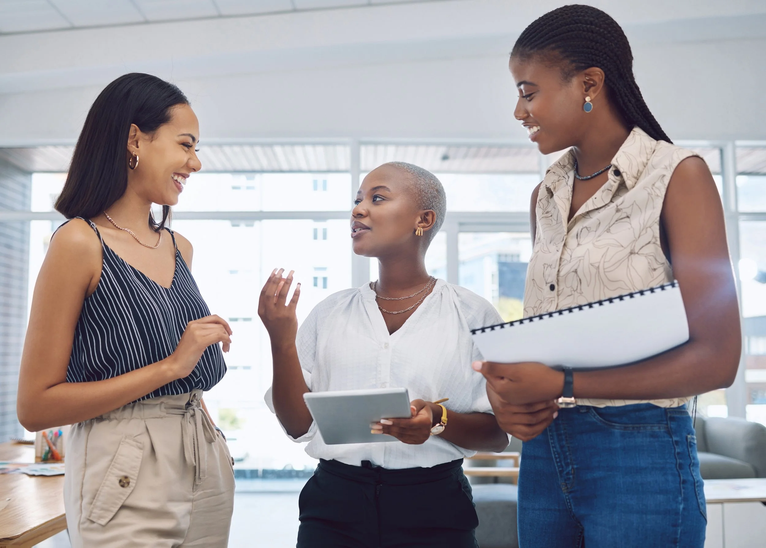 Three women having a conversation in a modern office, smiling and engaging with each other.