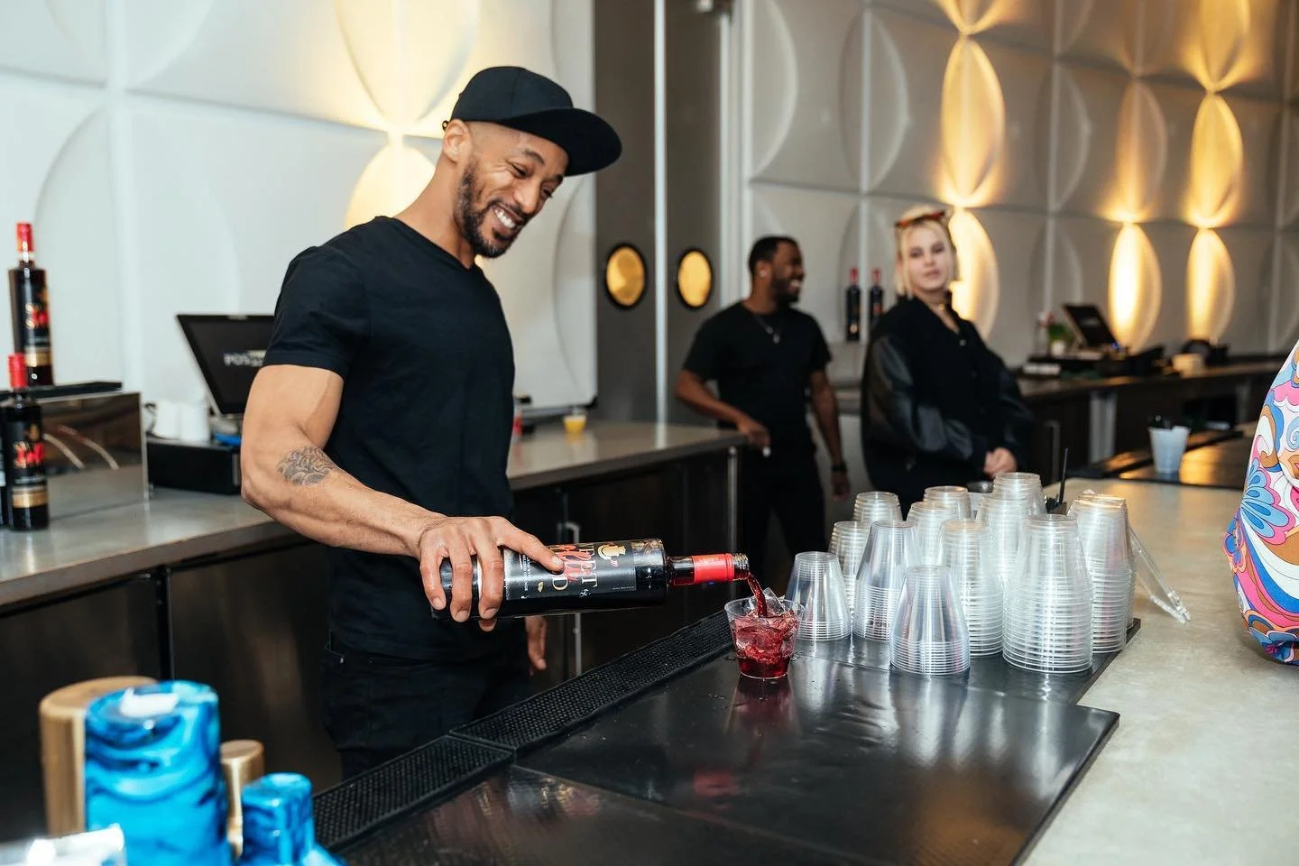 A man with a beard and tattoos wearing a black shirt and cap pours a red drink into a glass at a bar. Behind him, two other people stand by the bar.