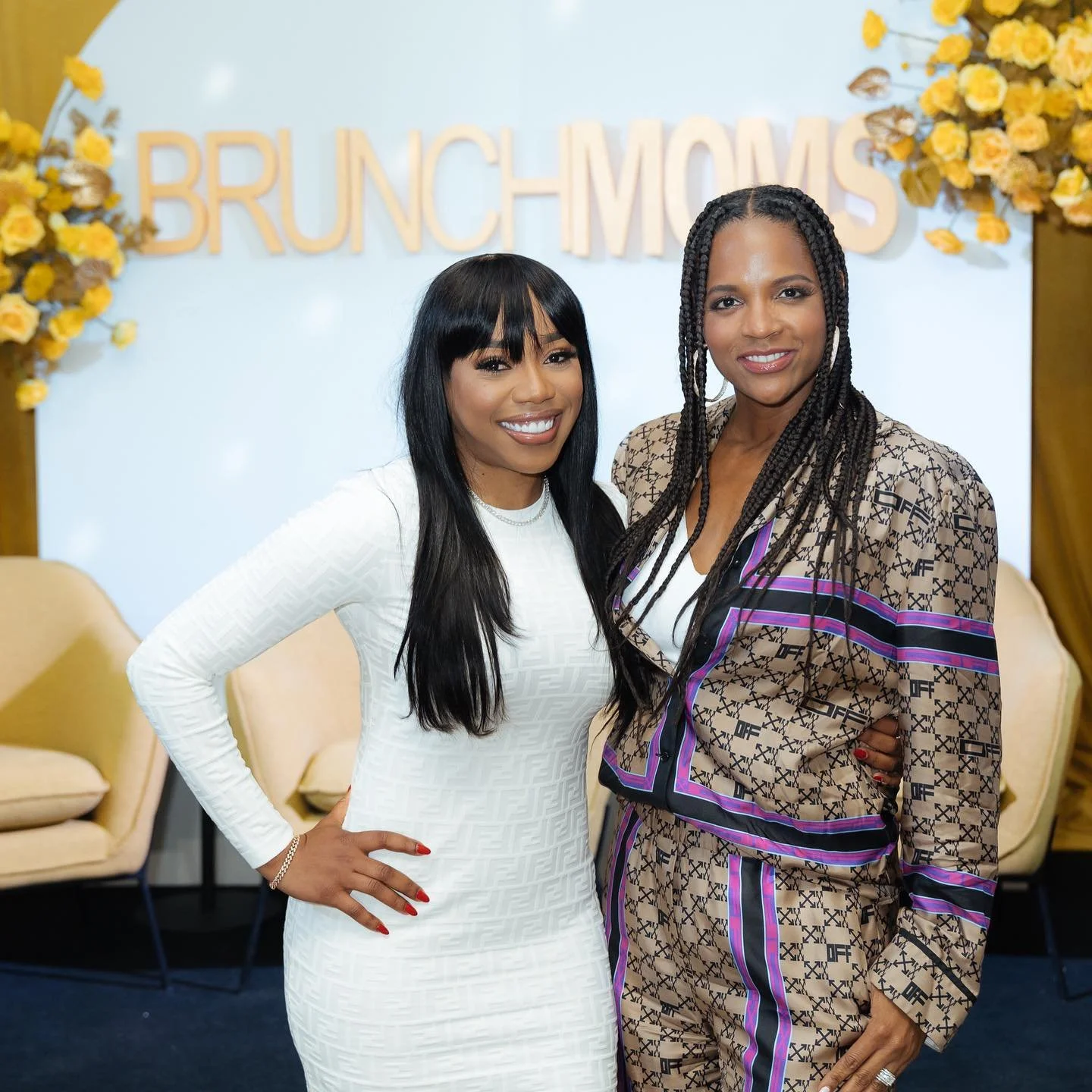 Two women smiling and standing close together in front of a floral backdrop with the words 'Brunch Mimosas'. One woman is wearing a white dress and the other is wearing a patterned outfit with braids.