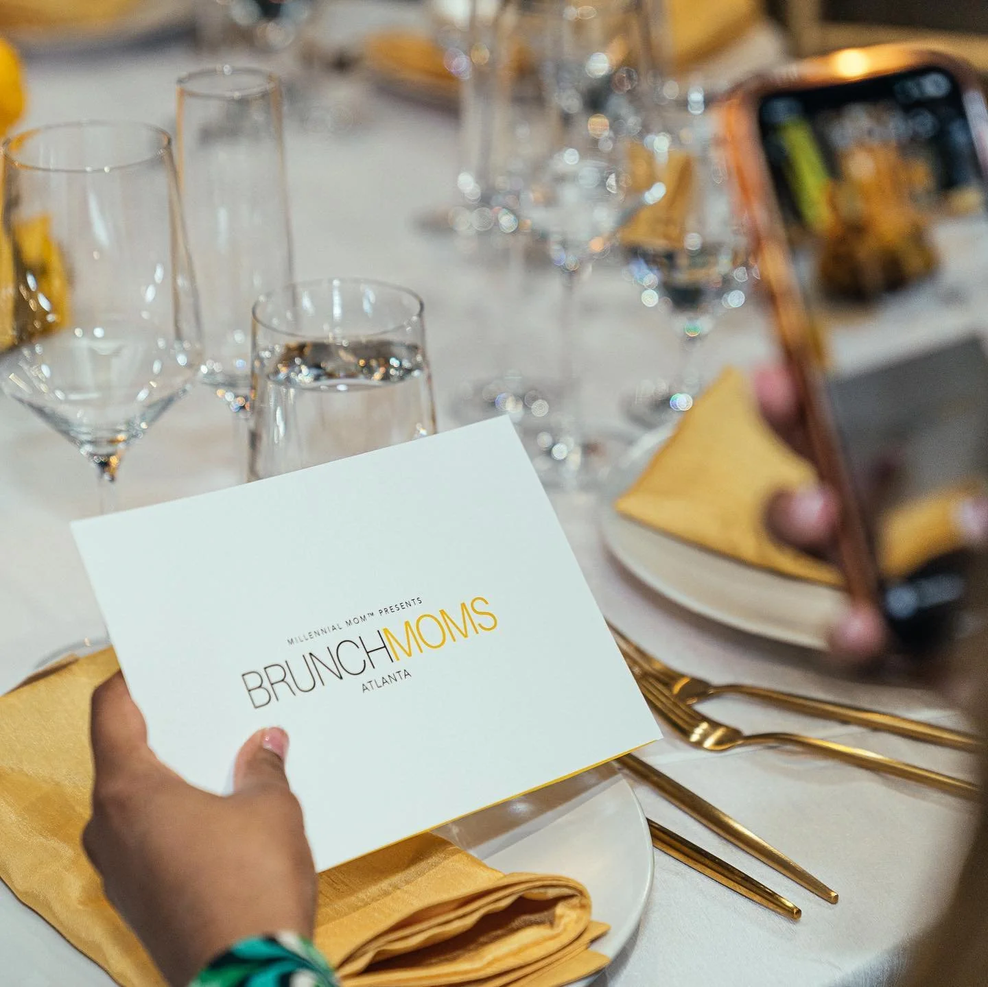 A person holding a card with the text 'Brunch Moms Atlanta' at a table set for a brunch event. The table has gold utensils, yellow napkins, glasses of water, and a plate with a folded yellow napkin. There is also a smartphone capturing the scene.
