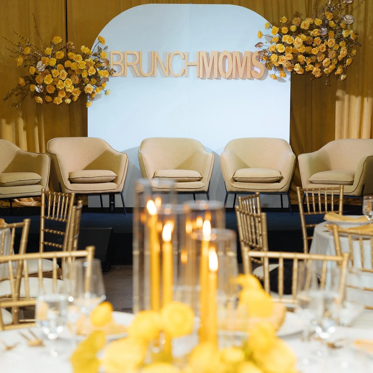 Stage setup with four beige armchairs, floral arrangements in yellow and purple, and a backdrop with 'Brunch Moms' sign, set for a brunch event.