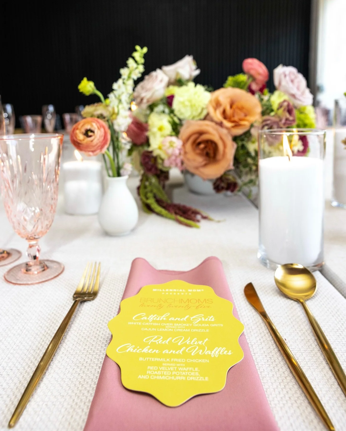Elegant table setting with a pink napkin, gold utensils, a menu card, a pink glass, and candles. In the background, there is a floral centerpiece with roses and other flowers in white and light pink shades.