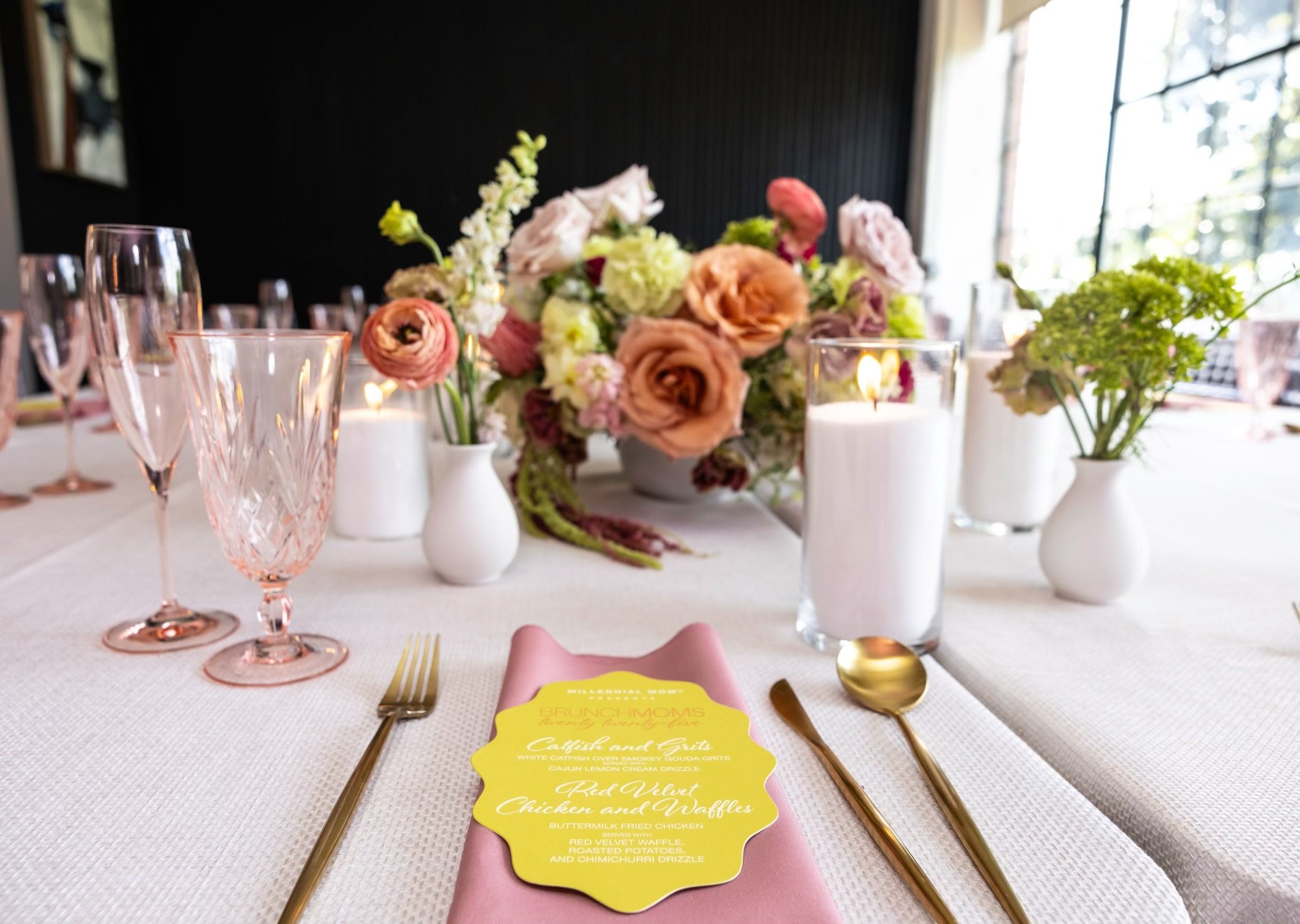 Elegant table setup with pink napkin, gold utensils, floral centerpiece, candles, and glassware for a special event or brunch.