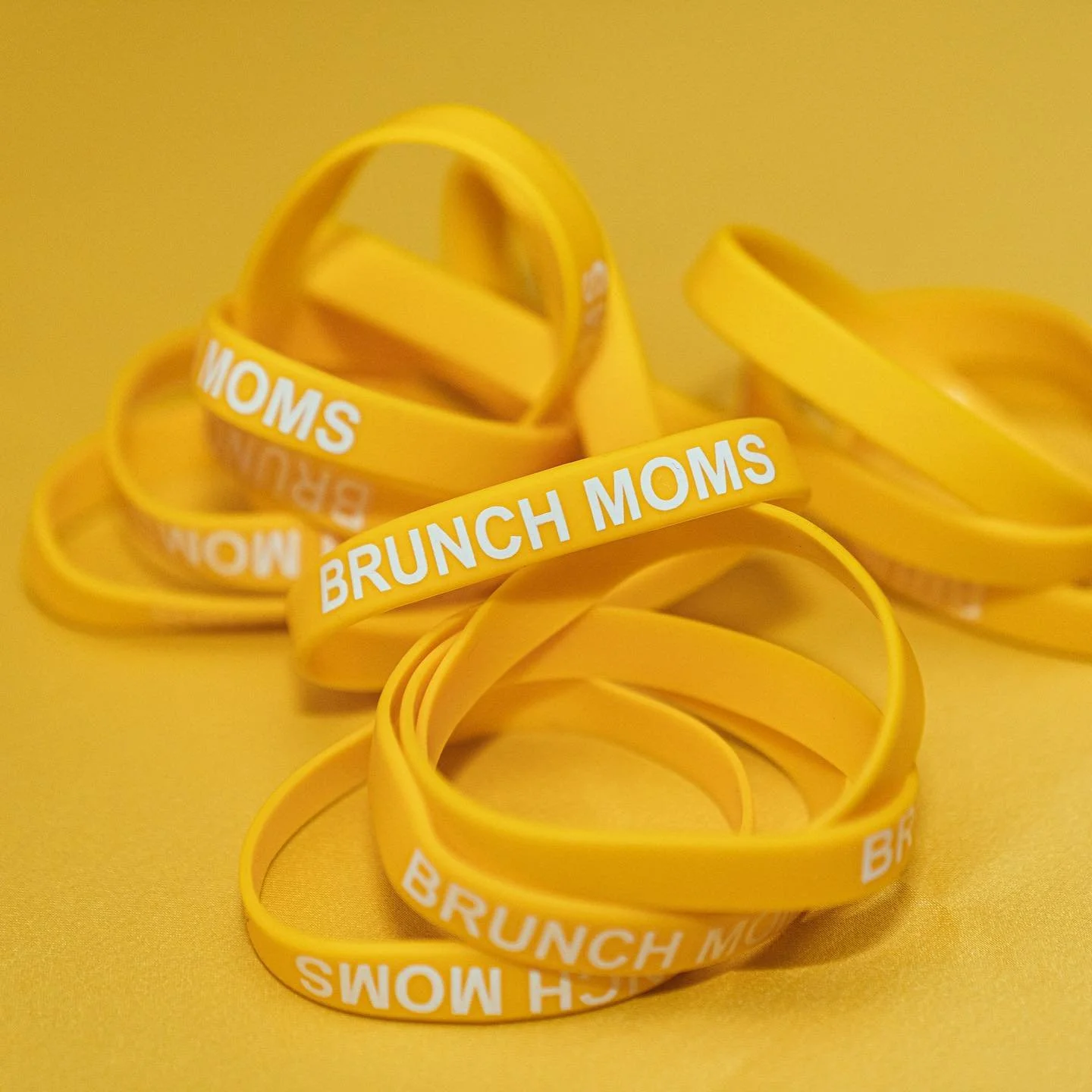 A collection of yellow silicone wristbands with the words 'BRUNCH MOMS' and 'MOMS' printed in white, arranged on a yellow background.