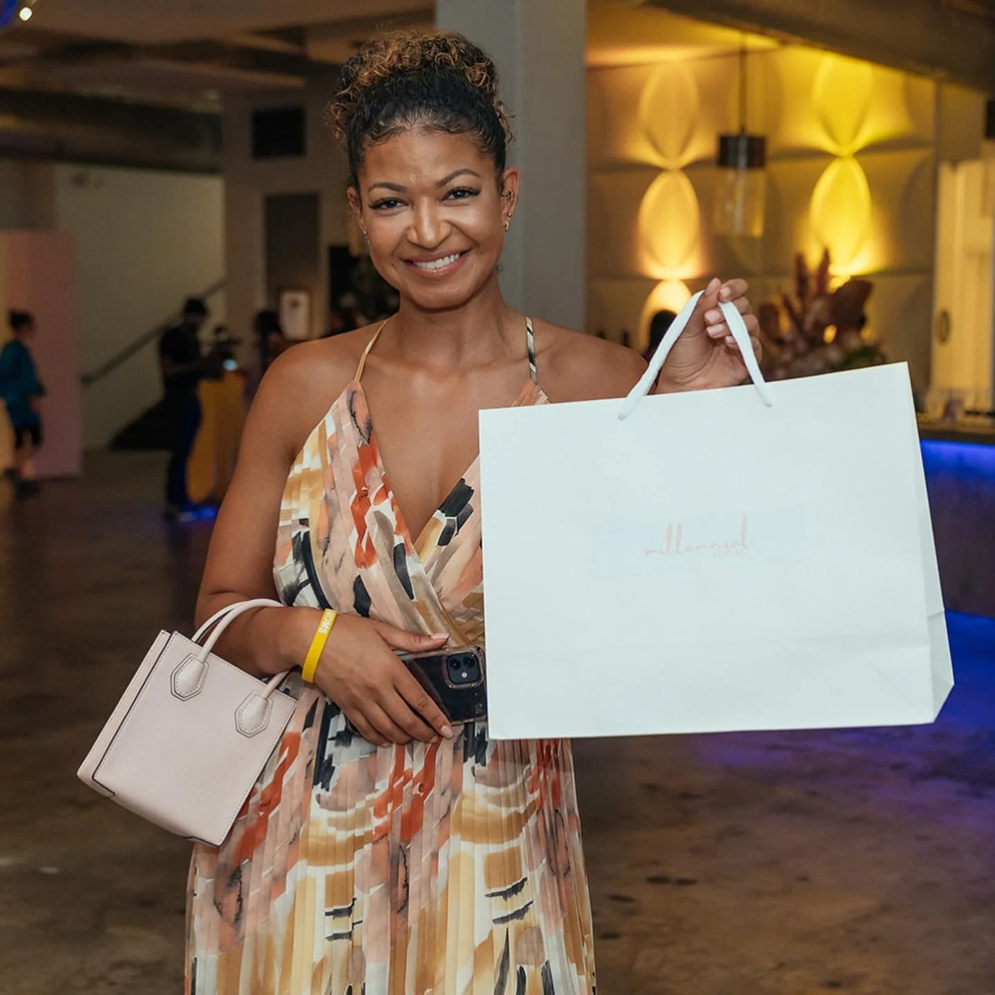A woman smiling and holding shopping bags and a smartphone in a lively indoor setting with warm lighting.