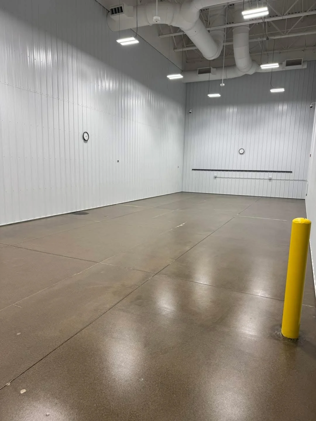 Empty industrial or commercial room with white paneled walls, a yellow safety post, and a concrete floor, with clocks on the walls and visible ceiling ducts.