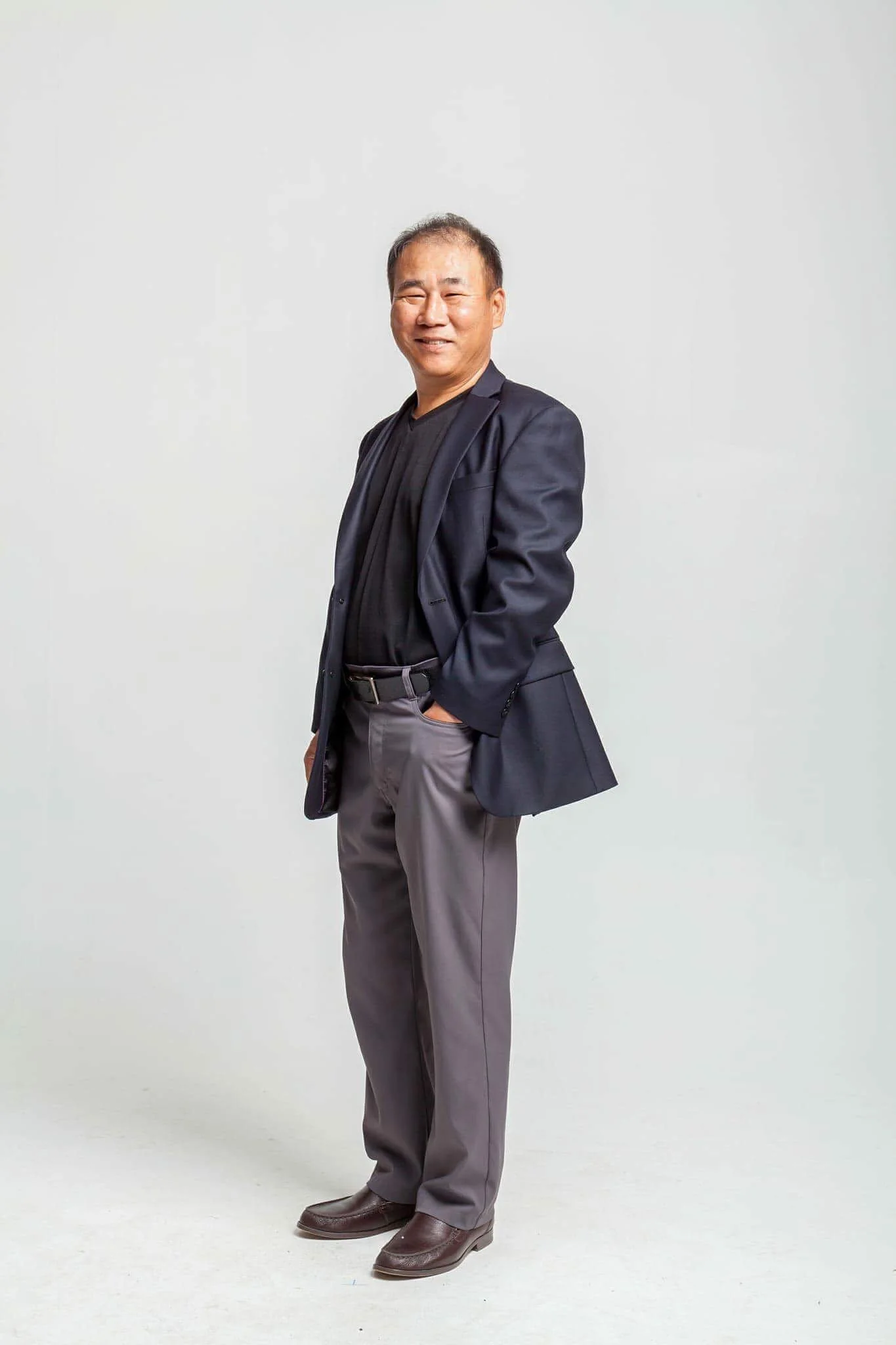 A man standing against a plain white background, smiling, wearing a dark blazer, black shirt, gray pants, and brown shoes, with his left hand in his pocket.
