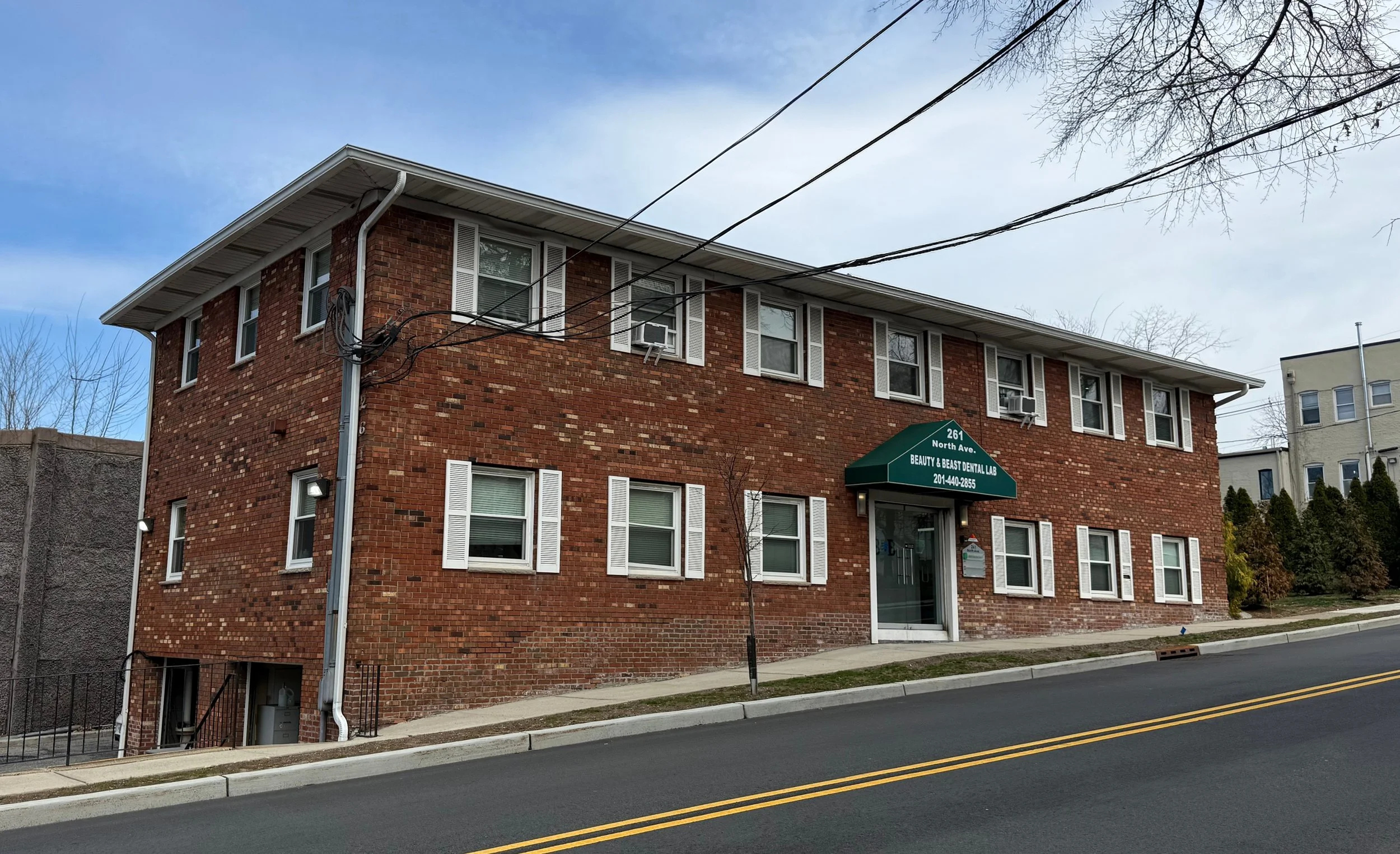 A three-story brick building with white window shutters, labeled as Beauty & Beast Dental Lab, with a green awning, located on a sloped street with a tree in front.