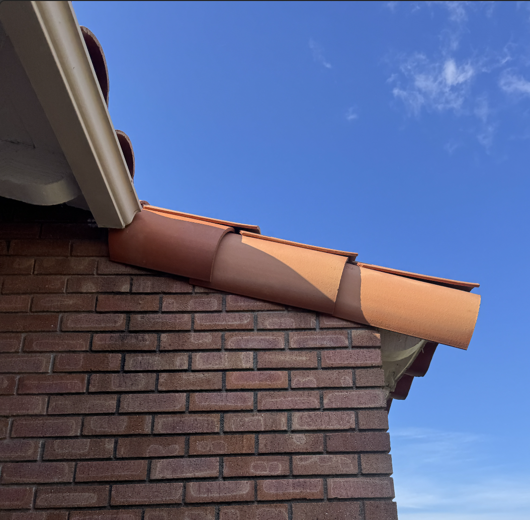 Close-up view of a brick house corner with terracotta roof tiles and a blue sky with some clouds.