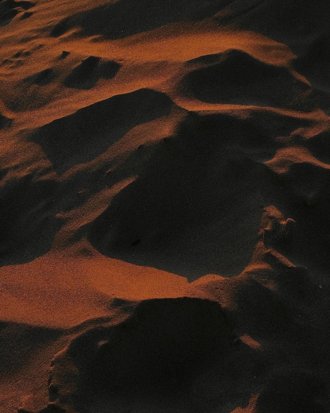 Close-up of sand dunes with wind-shaped patterns, illuminated by low or artificial light, creating shadows and highlights.