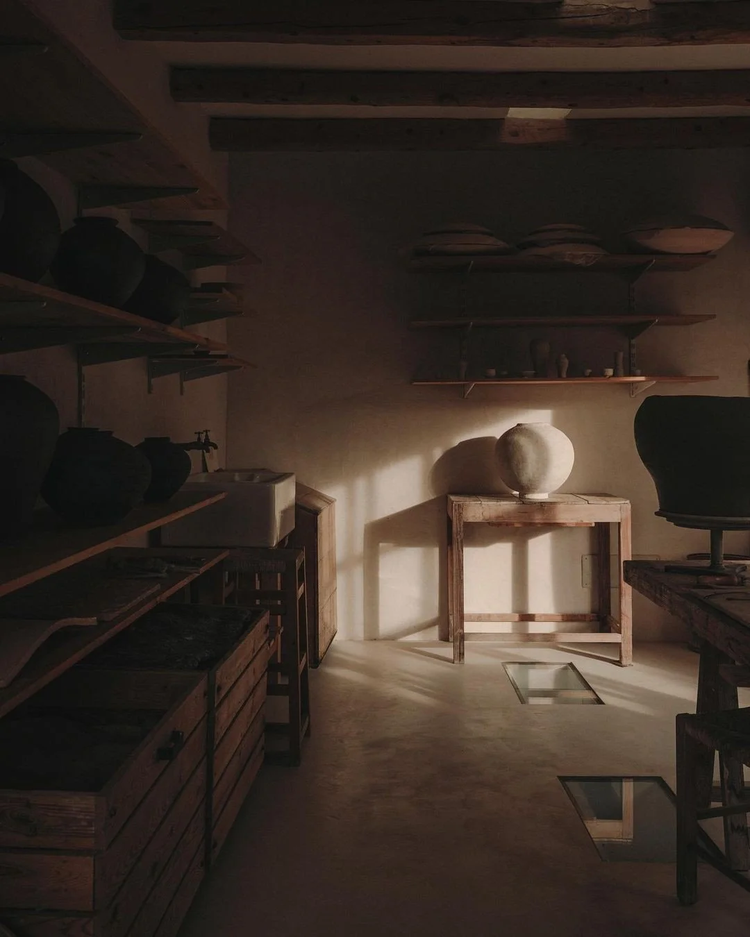 Darkly lit room with wooden shelves, crates, and tables displaying pottery and ceramic items, with sunlight creating shadows on the wall.