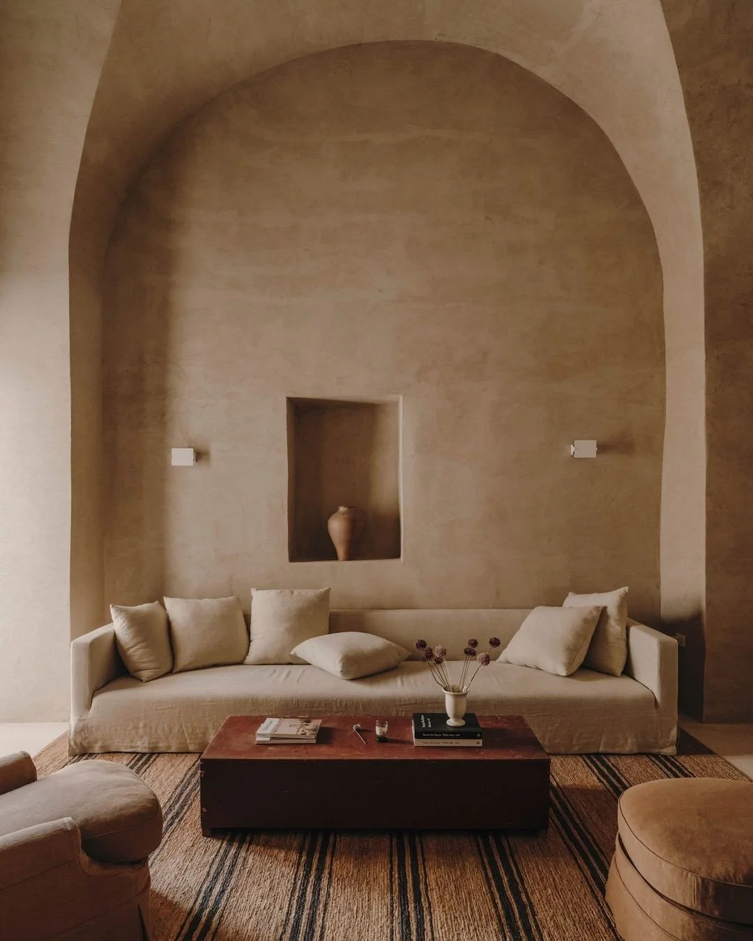 Minimalist living room with a beige sofa, several matching cushions, a wooden coffee table with magazines, a vase with dried flowers, a woven area rug, and a large arched wall opening with a decorative vase inside.