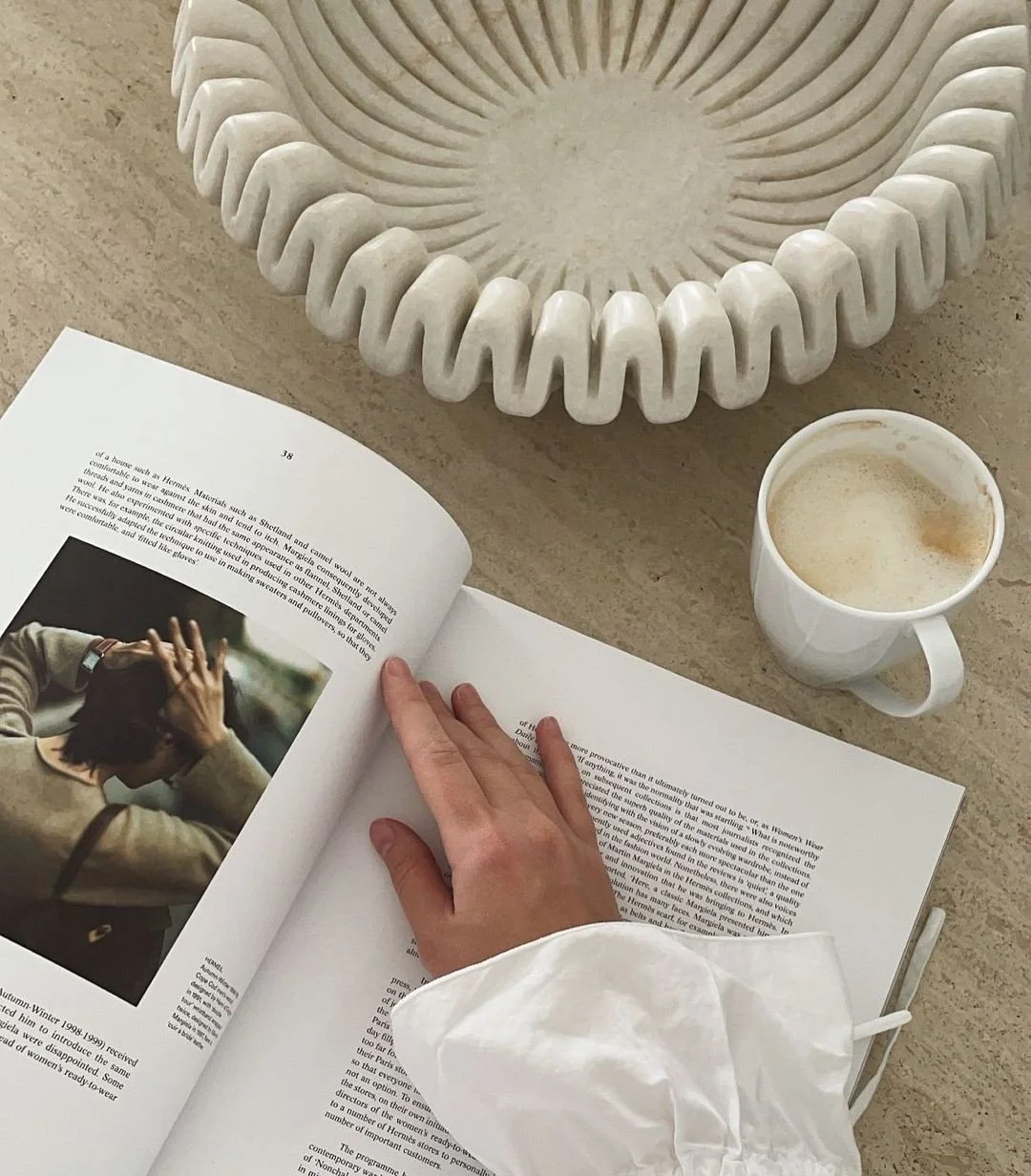 A magazine open on a table, a person’s hand flipping pages, a white coffee mug with coffee, and a decorative white tray with a wavy edge.