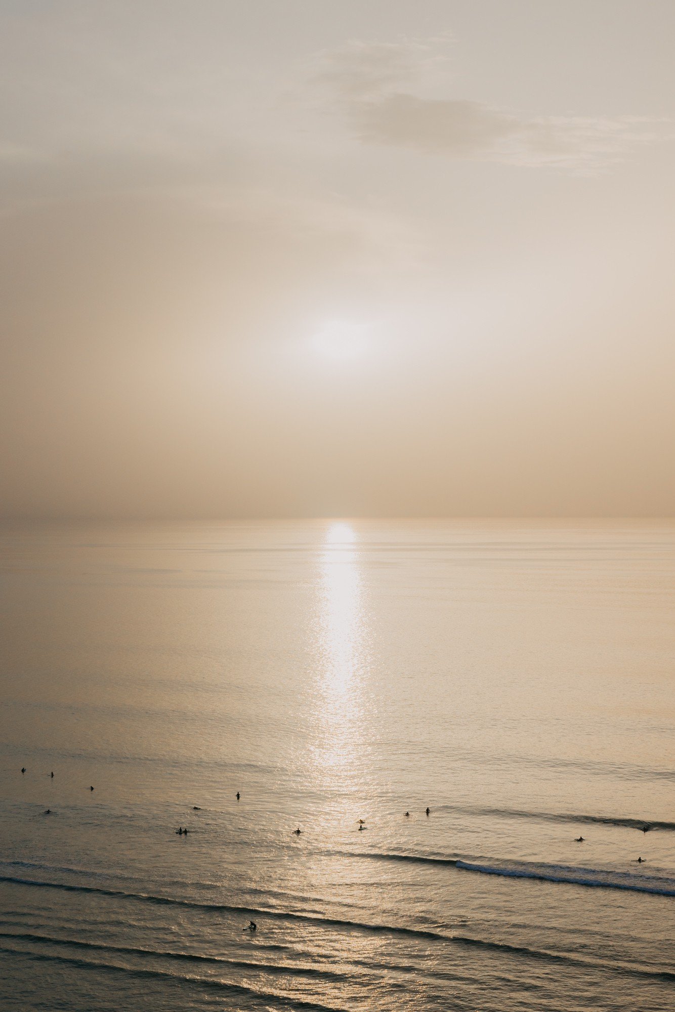 A serene seascape at sunset with a calm ocean, gentle waves, and a few silhouetted surfers in the water, under a sky with soft, muted colors.