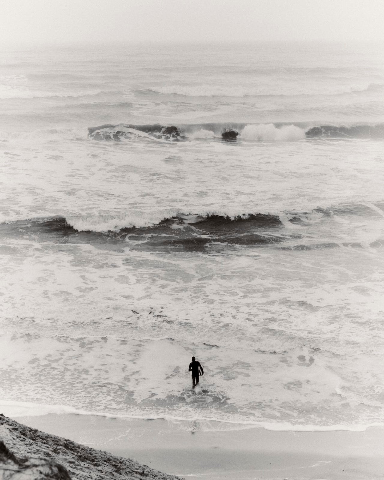 A person stands at the shoreline of a gray, overcast beach with waves crashing around them.
