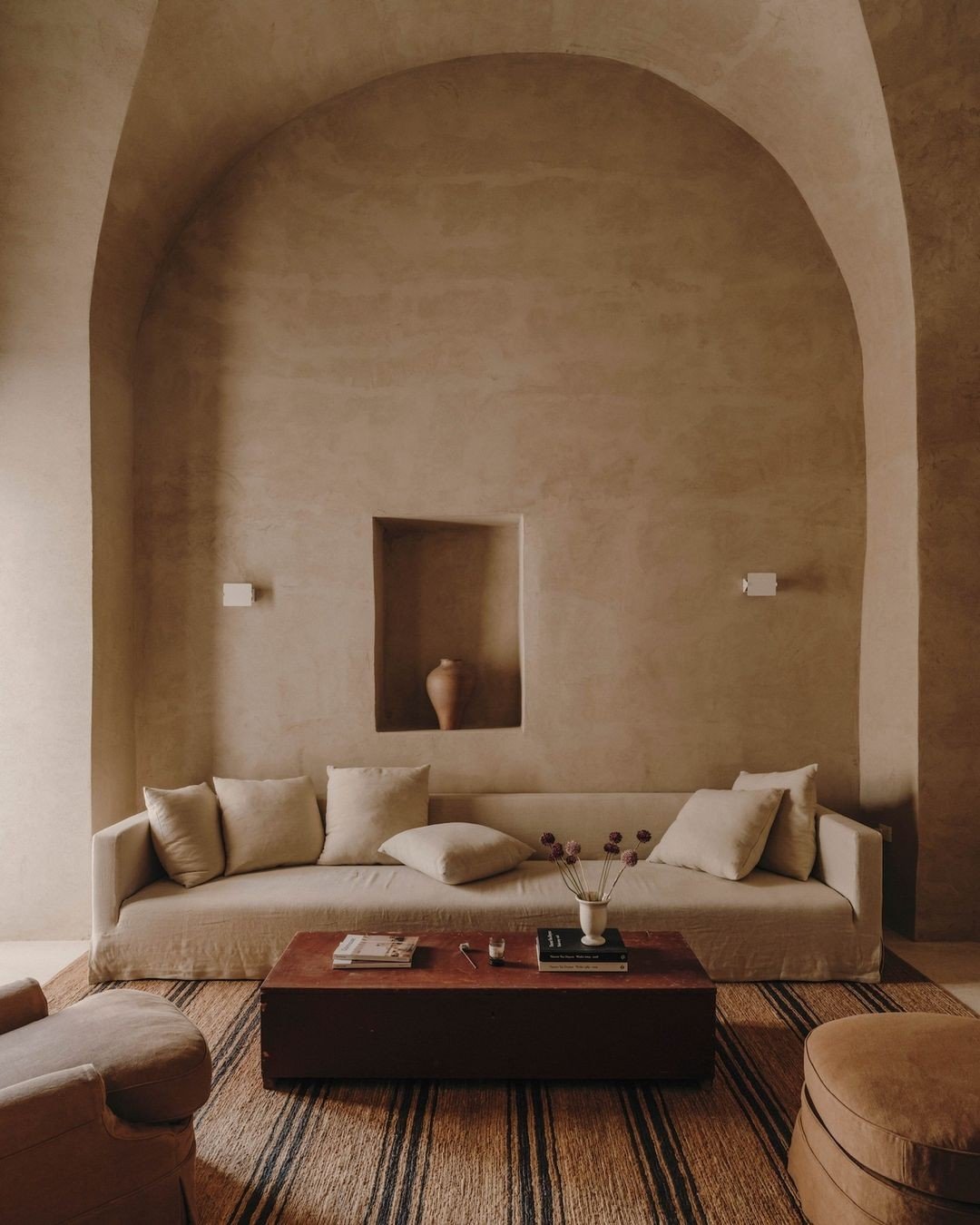Living room with a beige sofa, six beige pillows, a wooden coffee table with magazines, books, a small object, and a white vase with red flowers, against a textured beige wall with an arched ceiling and a recessed niche with a vase, and two wall sconces on either side.