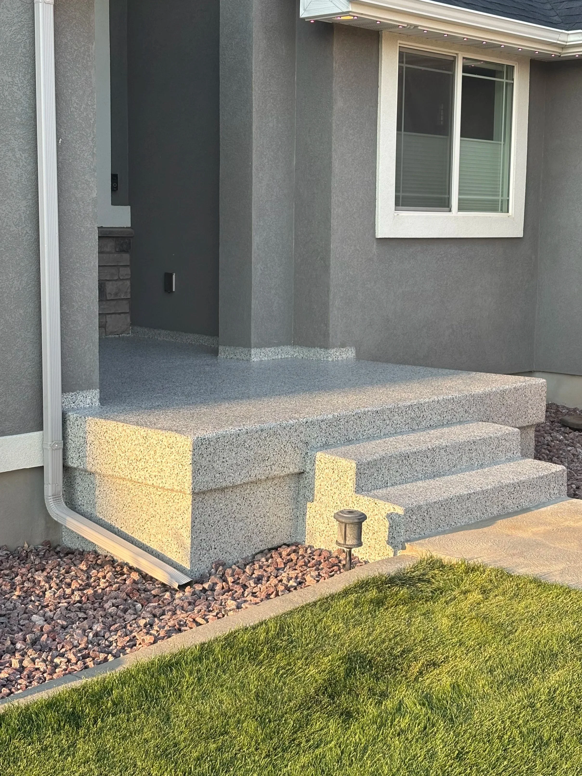 A front entrance with concrete steps, a small porch, and a gray exterior wall with a white-framed window, landscaping rocks, and grass in the yard.