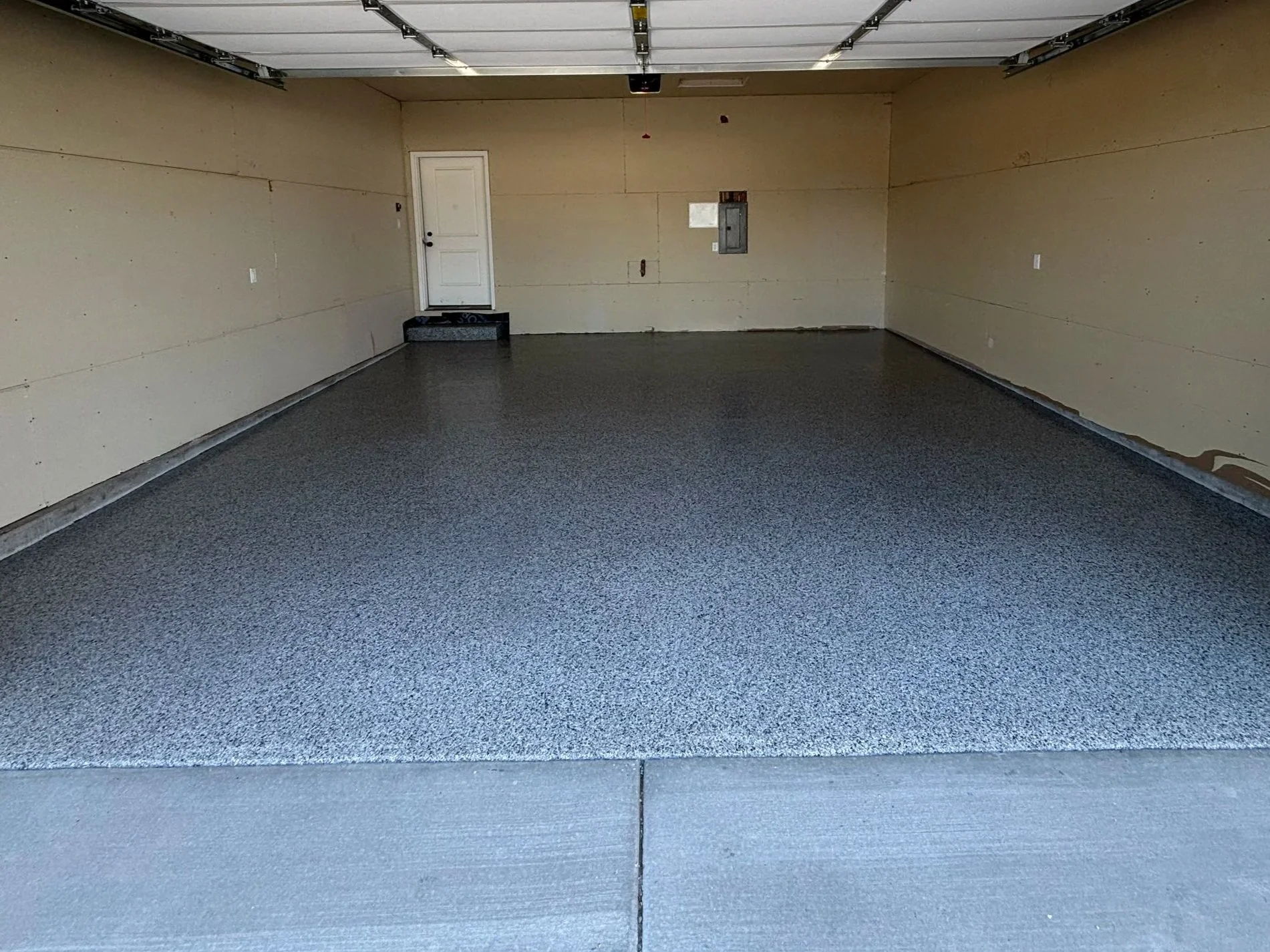 An empty two-car garage with a gray garage door, beige walls, gray carpeted flooring, a white door, and electrical panels on the wall.