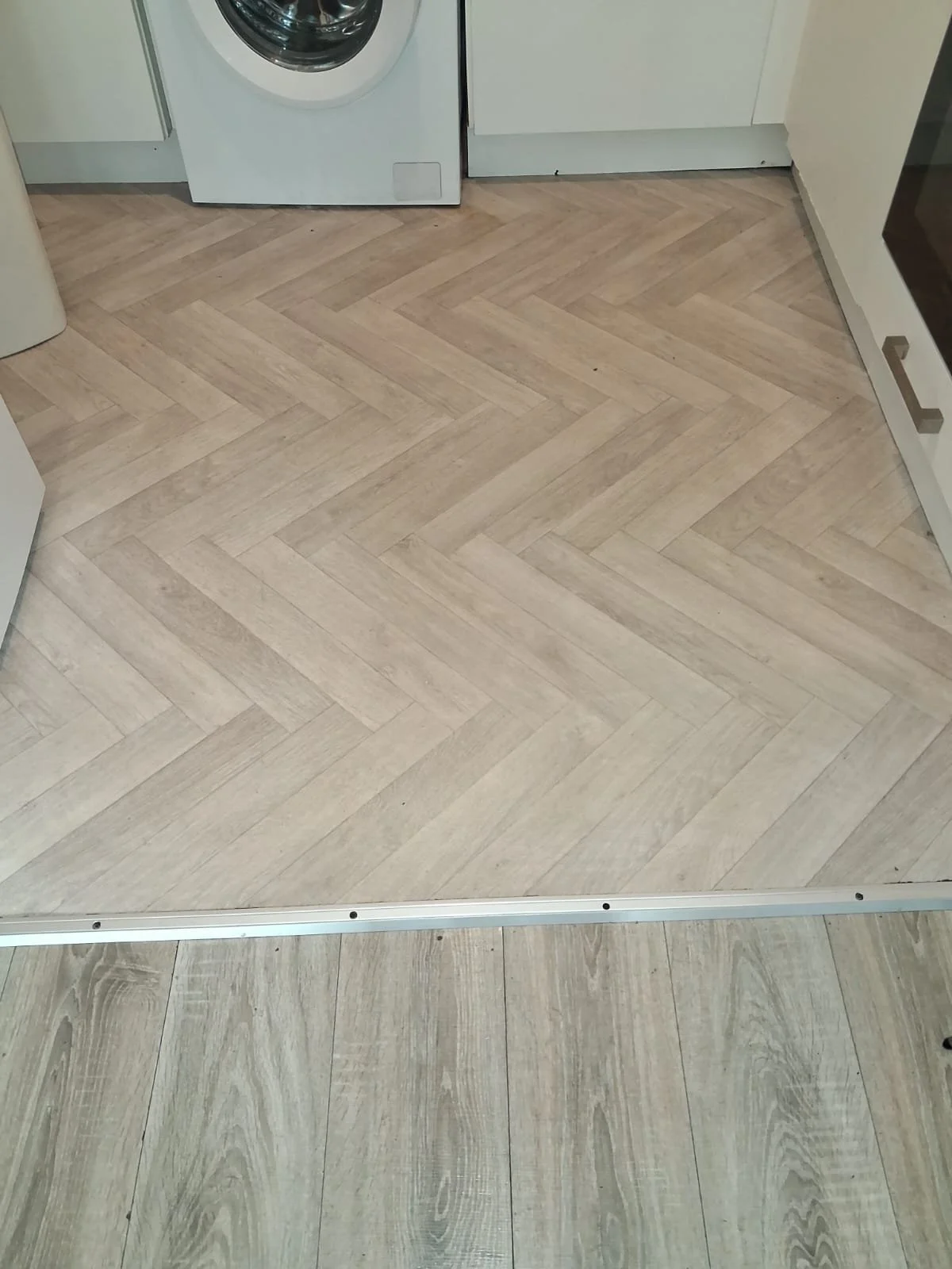 A kitchen floor with two types of light-colored wood laminate flooring, separated by a metallic strip. In the background, a washing machine is partially visible, along with white kitchen cabinets.