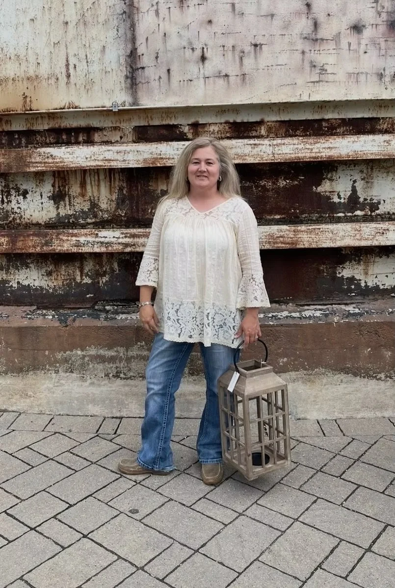 A woman with blonde hair in a white lace top, blue jeans, and brown boots stands on a cobblestone sidewalk holding a gray wooden lantern, with a rusty metal wall behind her.