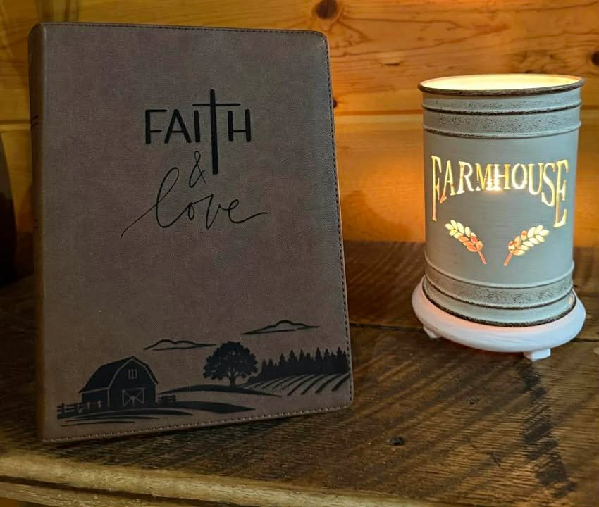 A gray notebook with the words 'Faith & Love' on the cover, and a decorative illustration of a barn, tree, and mountains. Next to it is a candle labeled 'Farmhouse' on a wooden surface.