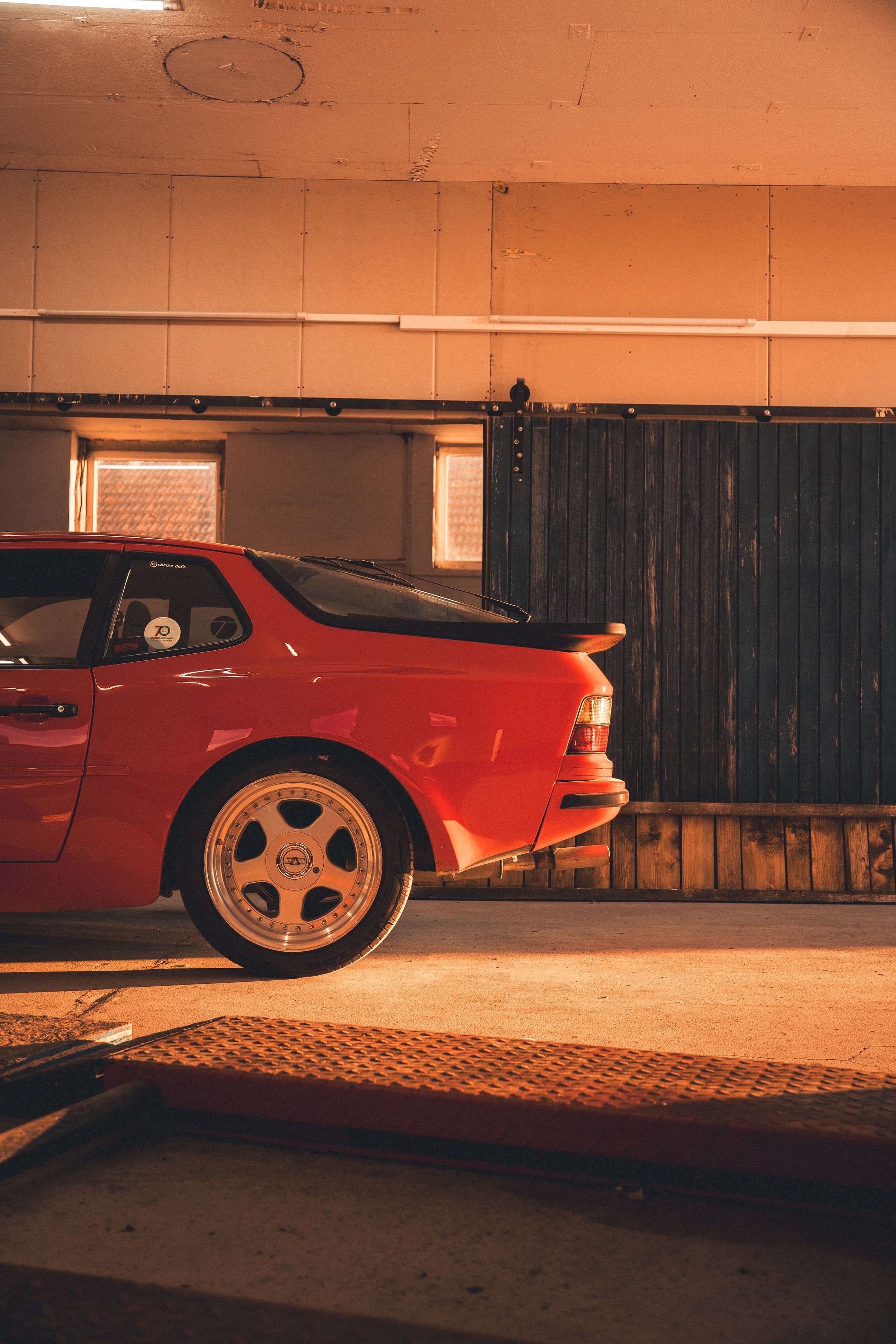 Ein rotes Auto steht in einer Garage mit Holz- und Trockenbauwänden, nur die Rückseite des Autos ist sichtbar, mit modernen Felgen und einem Spoiler.