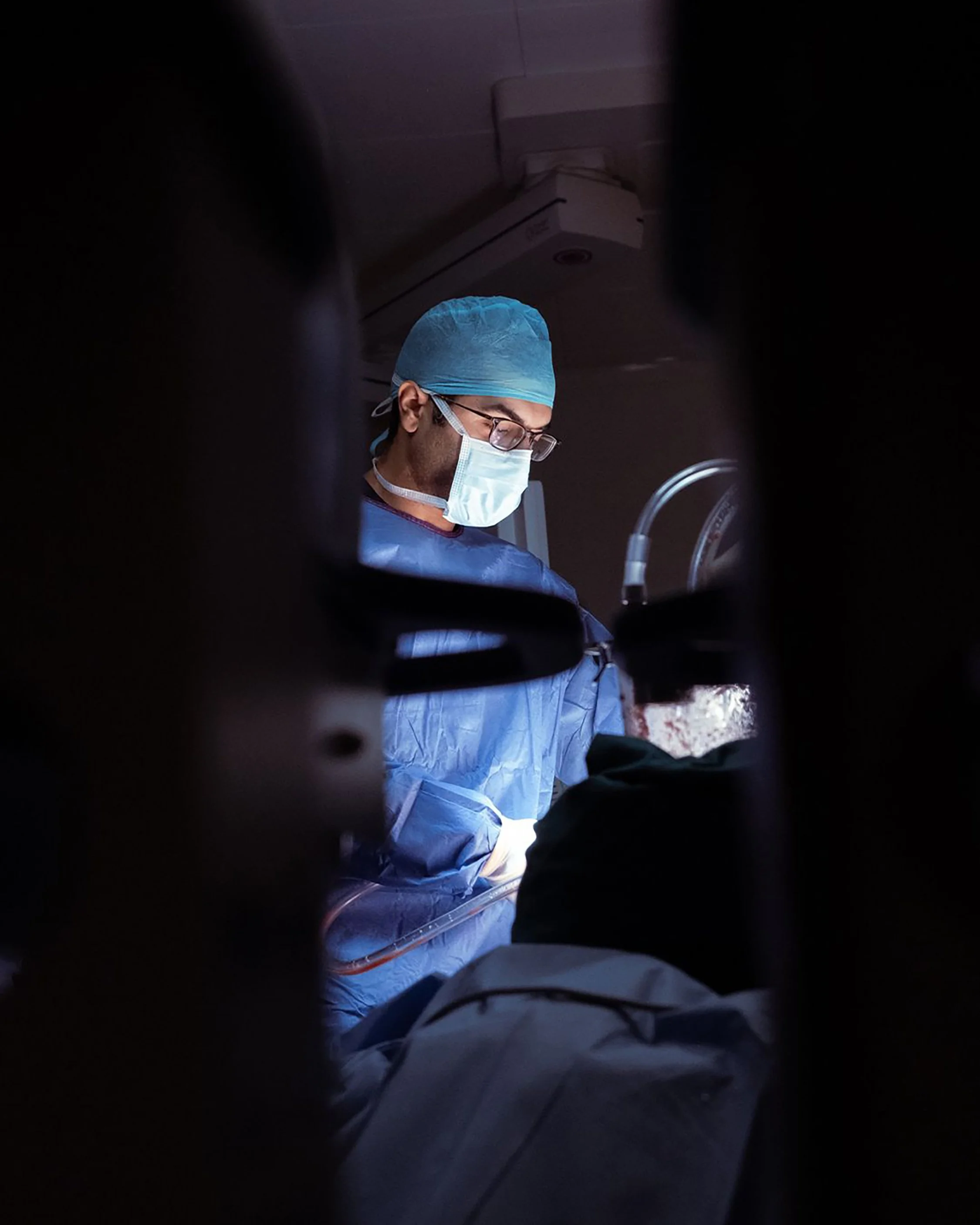Medical professional wearing a blue surgical cap, face mask, and glasses working in a dimly lit operating room.