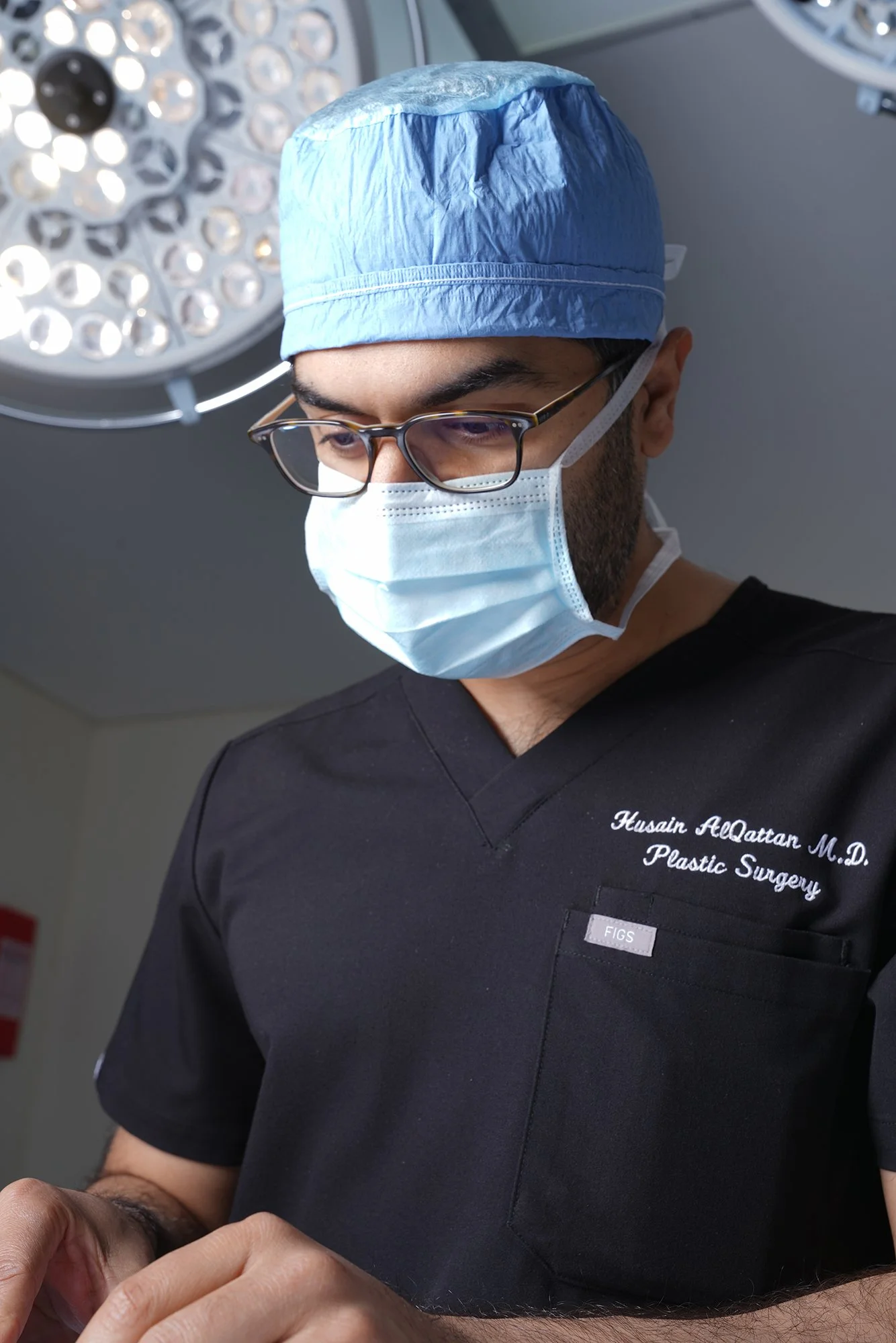 A male surgeon wearing a scrub cap, face mask, glasses, and black scrub top with name tag, in an operating room with surgical lights.