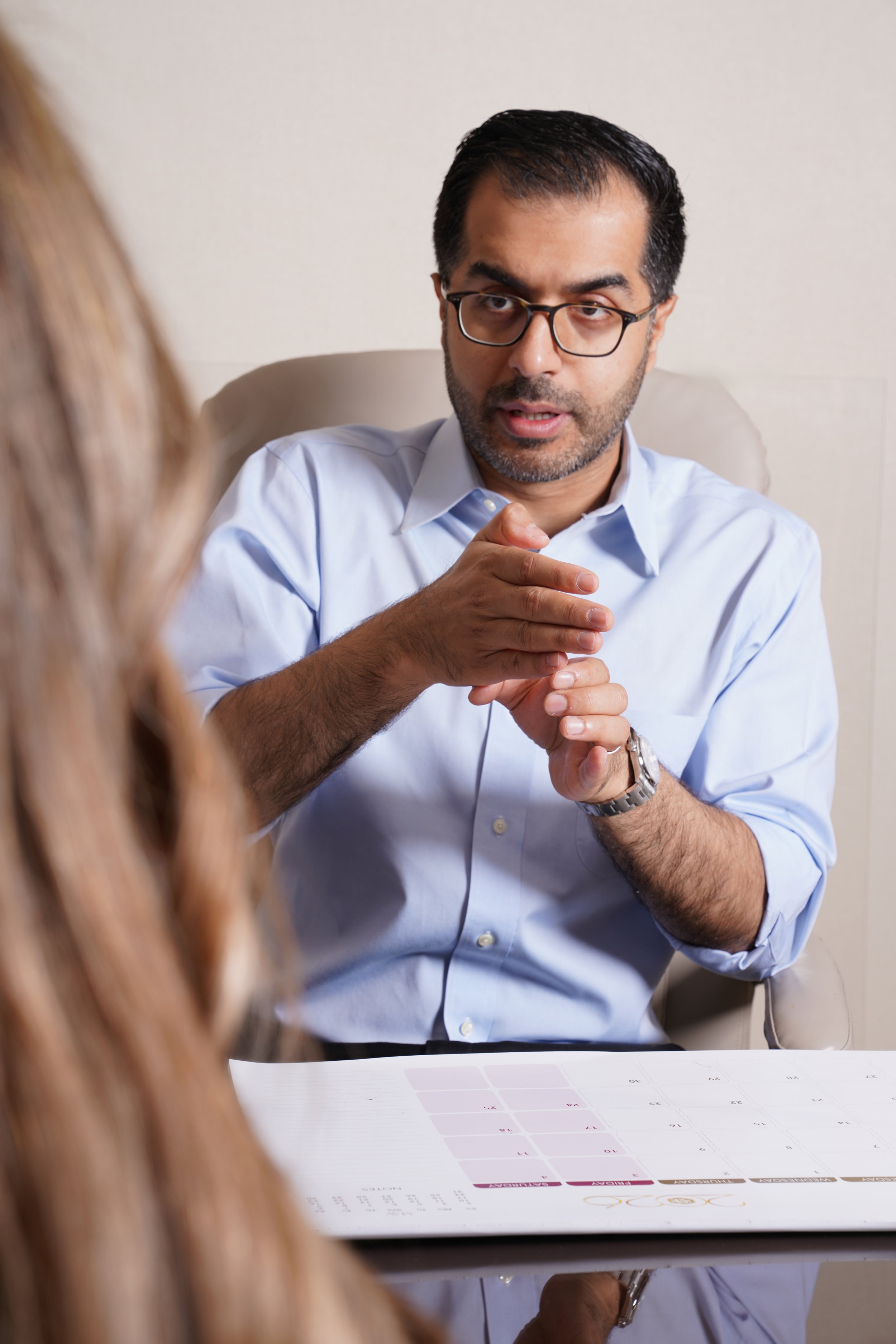 Man wearing glasses and a light blue shirt speaking during a consultation or meeting, with a woman partially visible in the foreground, sitting at a table with a printed calendar or schedule.