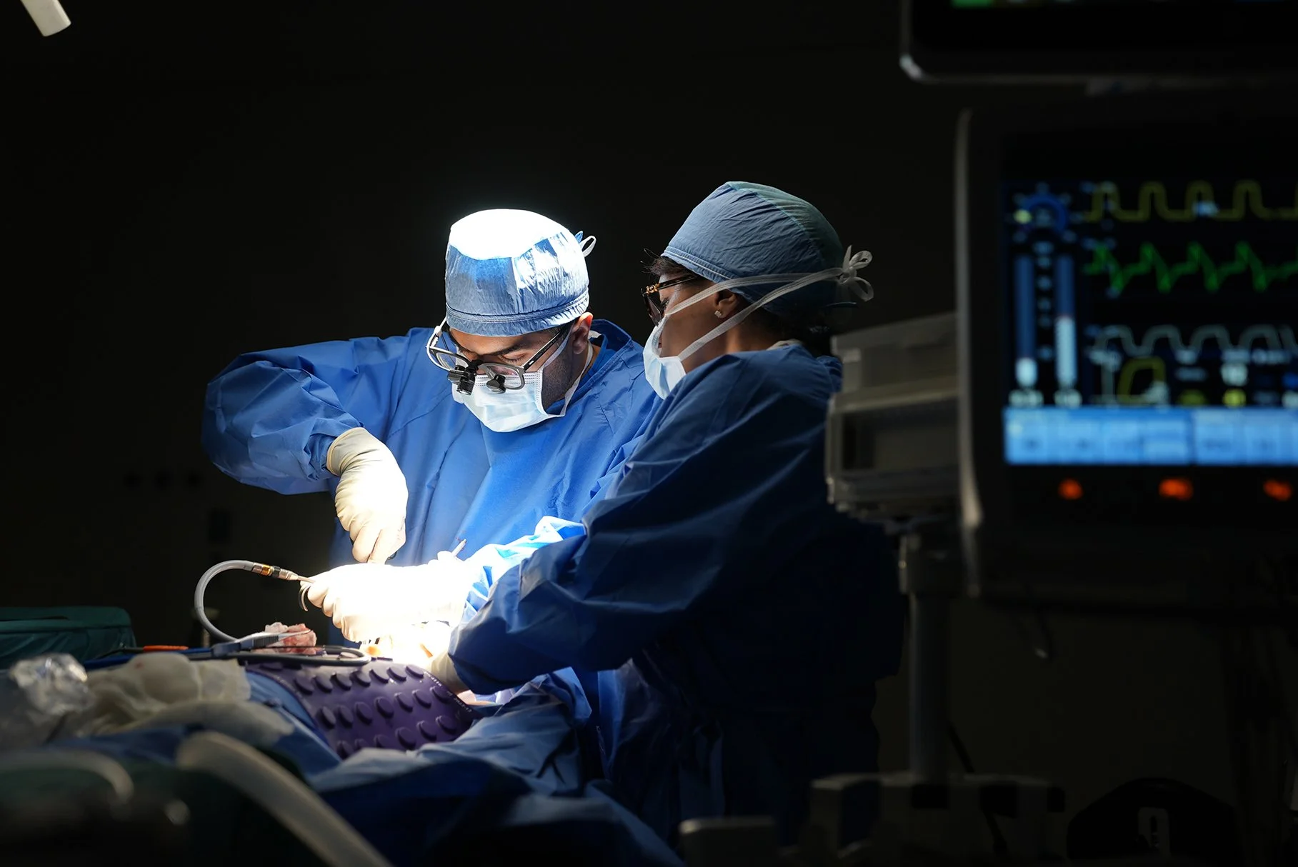 Two surgeons wearing blue scrubs, masks, and surgical caps performing an operation in a dimly lit operating room, with a medical monitor displaying vital signs nearby.