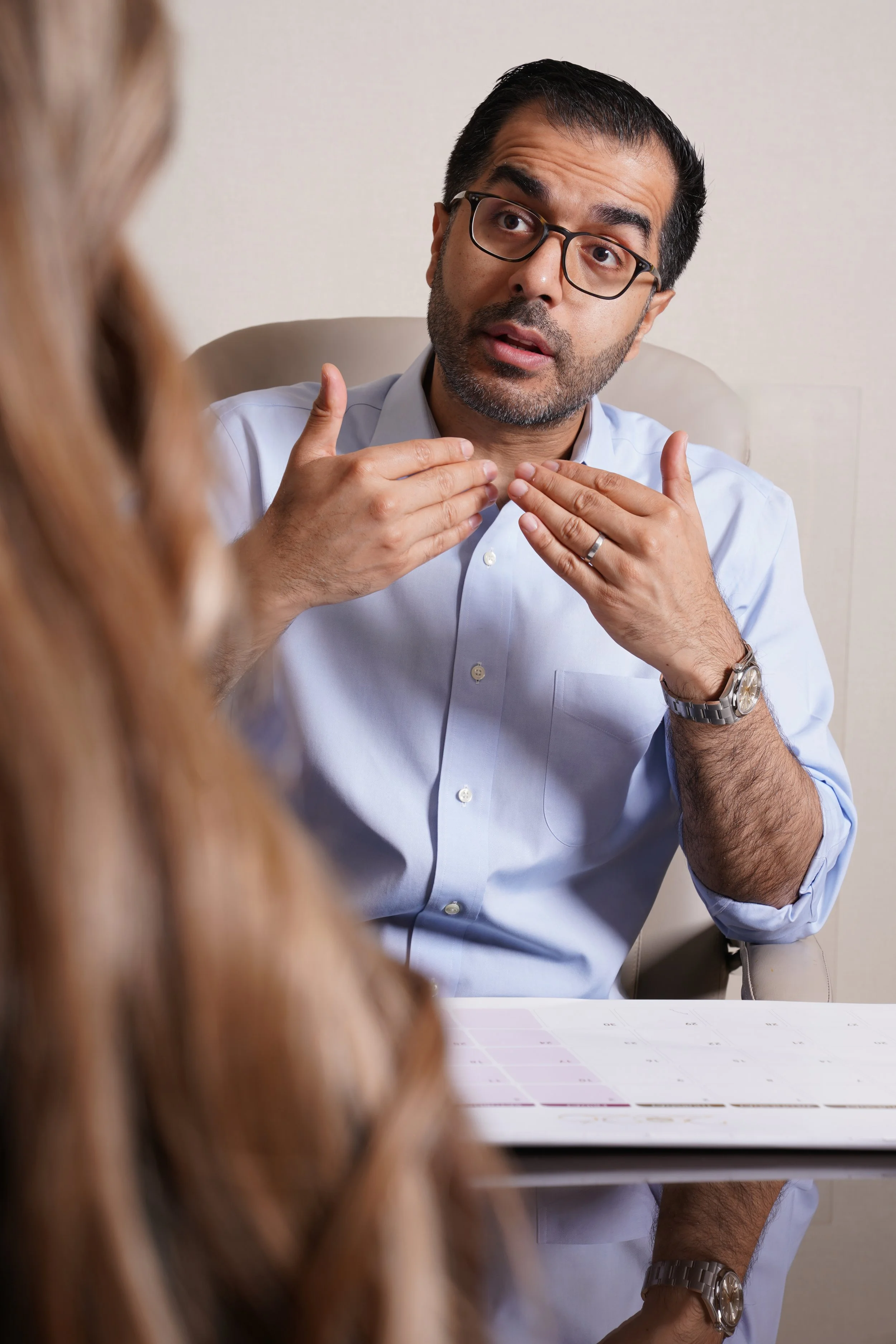 A man with glasses and a beard, wearing a light blue shirt, is speaking and gesturing with his hands during a conversation, sitting at a desk with a chart or calendar in front of him. A woman with long, wavy blonde hair is visible in the foreground, blurred out.